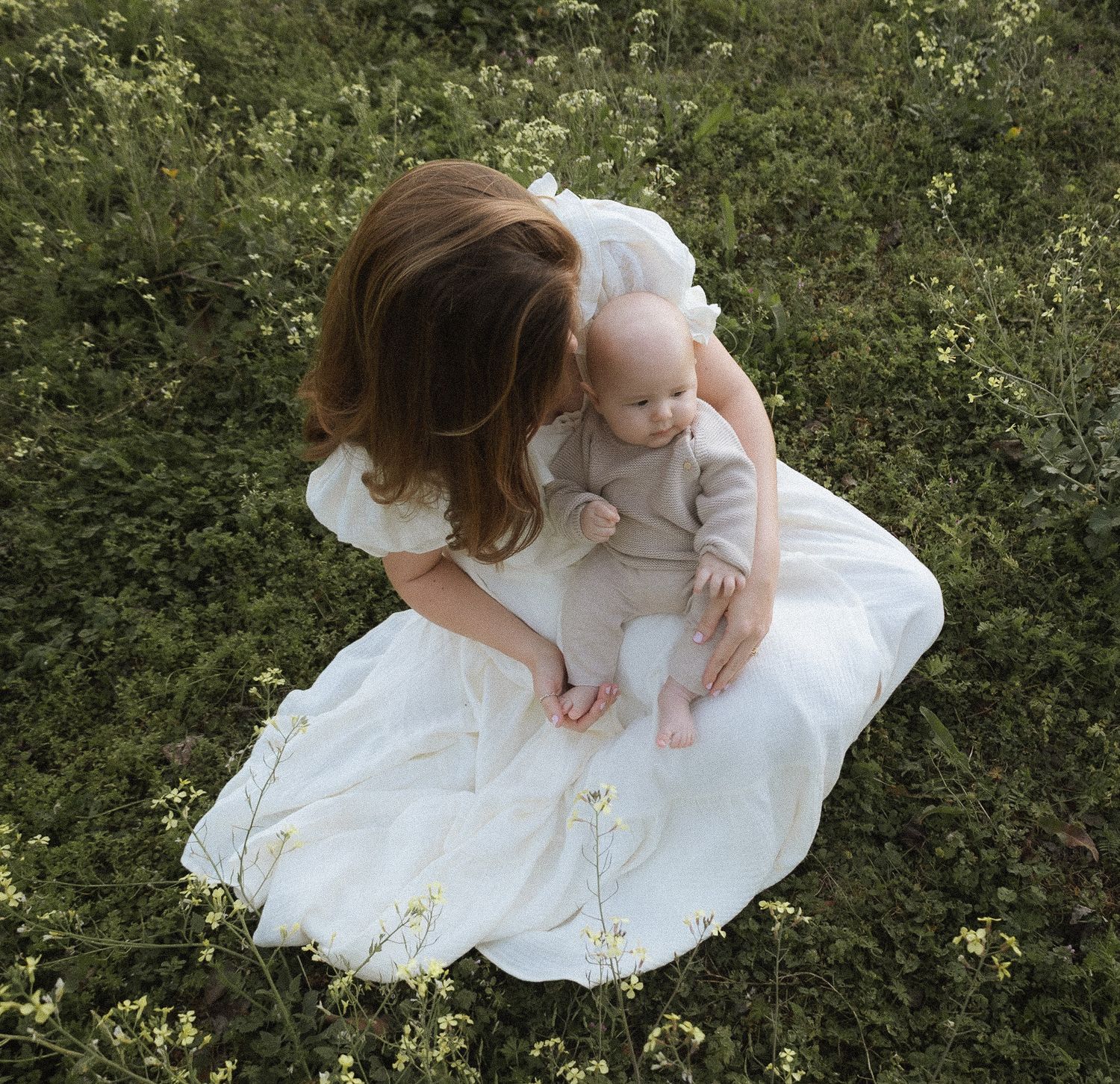 Overhead view of mother in white dress sitting in wildflower meadow with baby.
