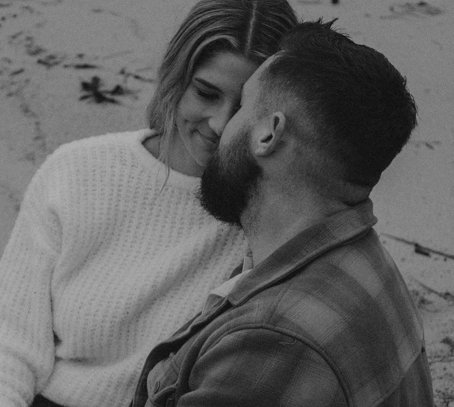 Black and white close-up of a bearded man and smiling woman sharing an intimate moment on a sandy beach.