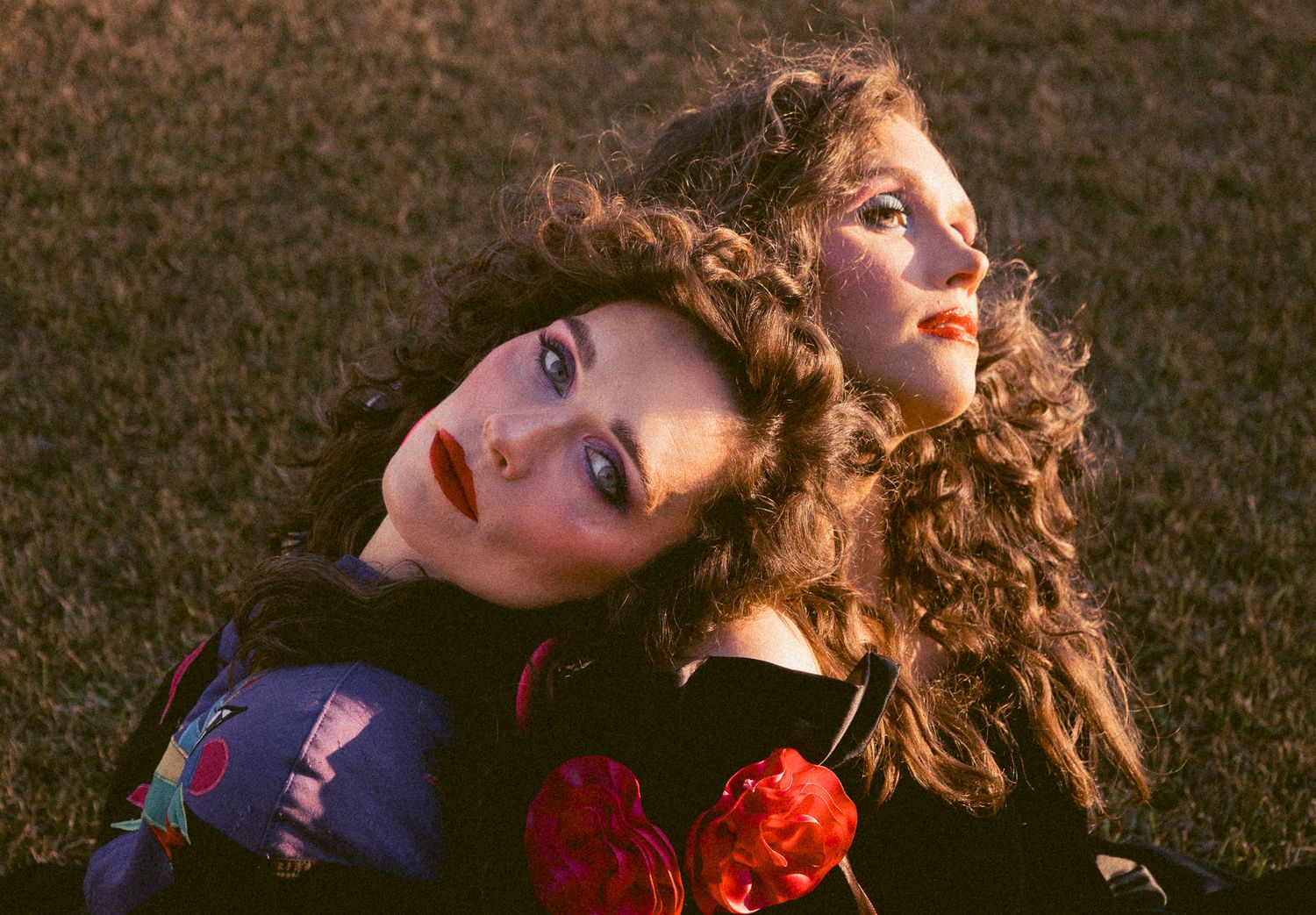 Two women with curly hair and red lips lying on grass together in colorful vintage outfits at sunset.