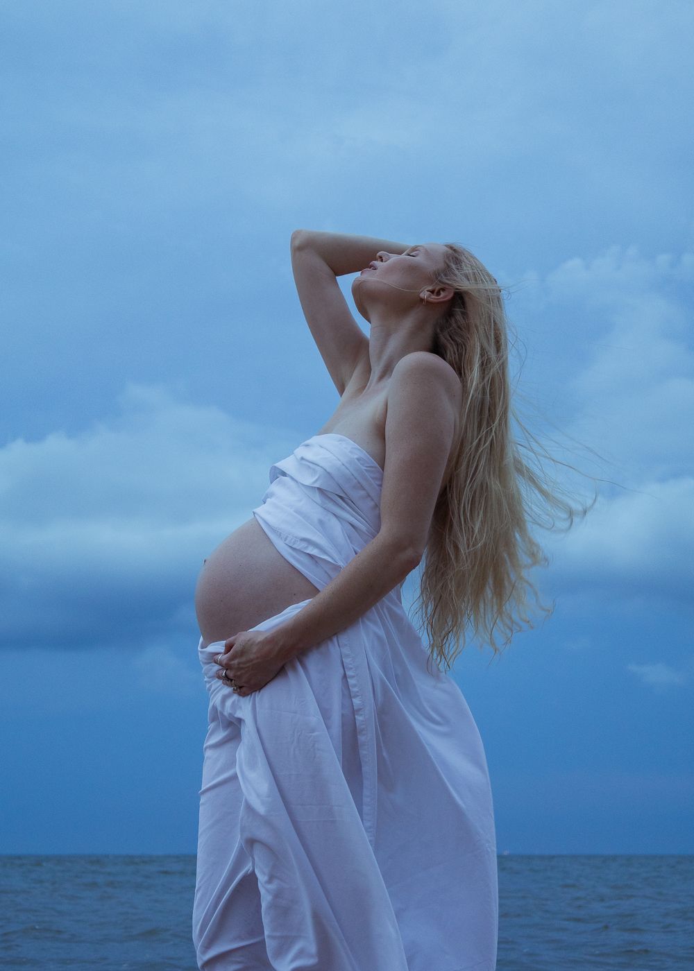 Pregnant woman in white dress stands by the ocean, wind in her blonde hair, against a dramatic blue cloudy sky.