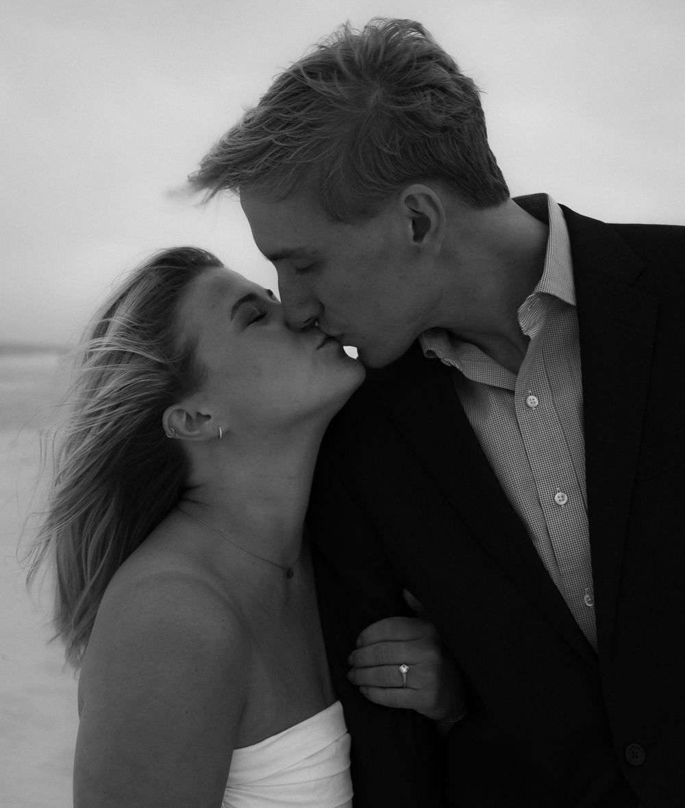 Couple shares a romantic kiss on the beach in a classic black and white engagement photo.