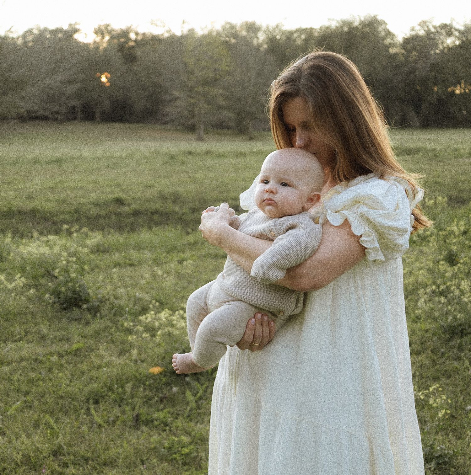 Tender mother in white dress kissing newborn baby while standing in sunlit green field at dusk.
