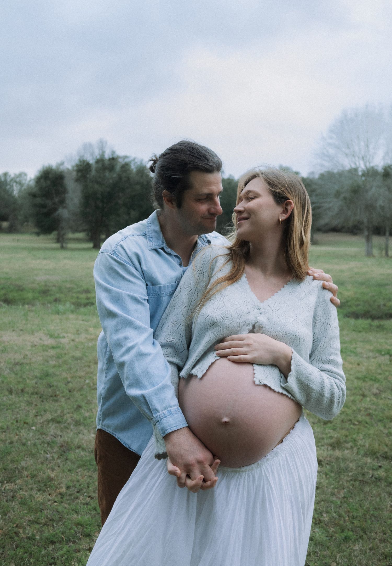 Happy couple embracing in green field, pregnant woman in white dress with exposed baby bump.