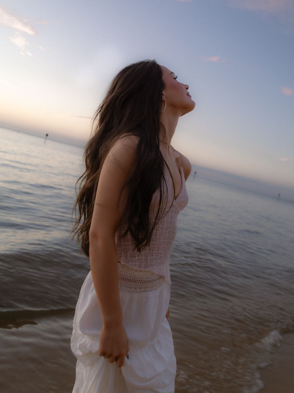 Woman in white dress at beach sunset, hair flowing, head tilted back enjoying the ocean breeze.