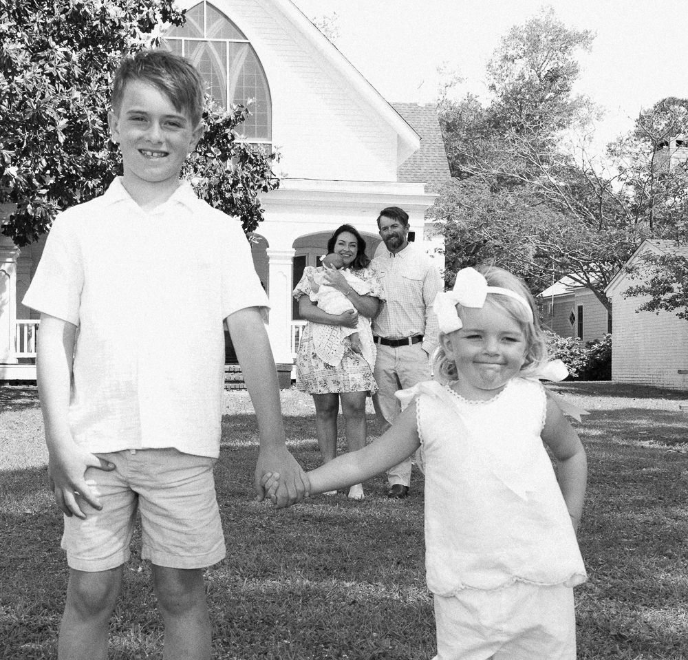 Family posing outdoors near a white church, two children holding hands in foreground, parents with baby in background.