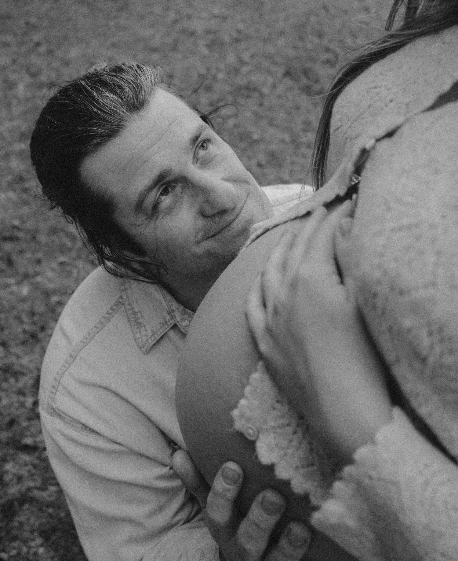 Black and white close-up of man looking up while embracing pregnant partner's baby bump.