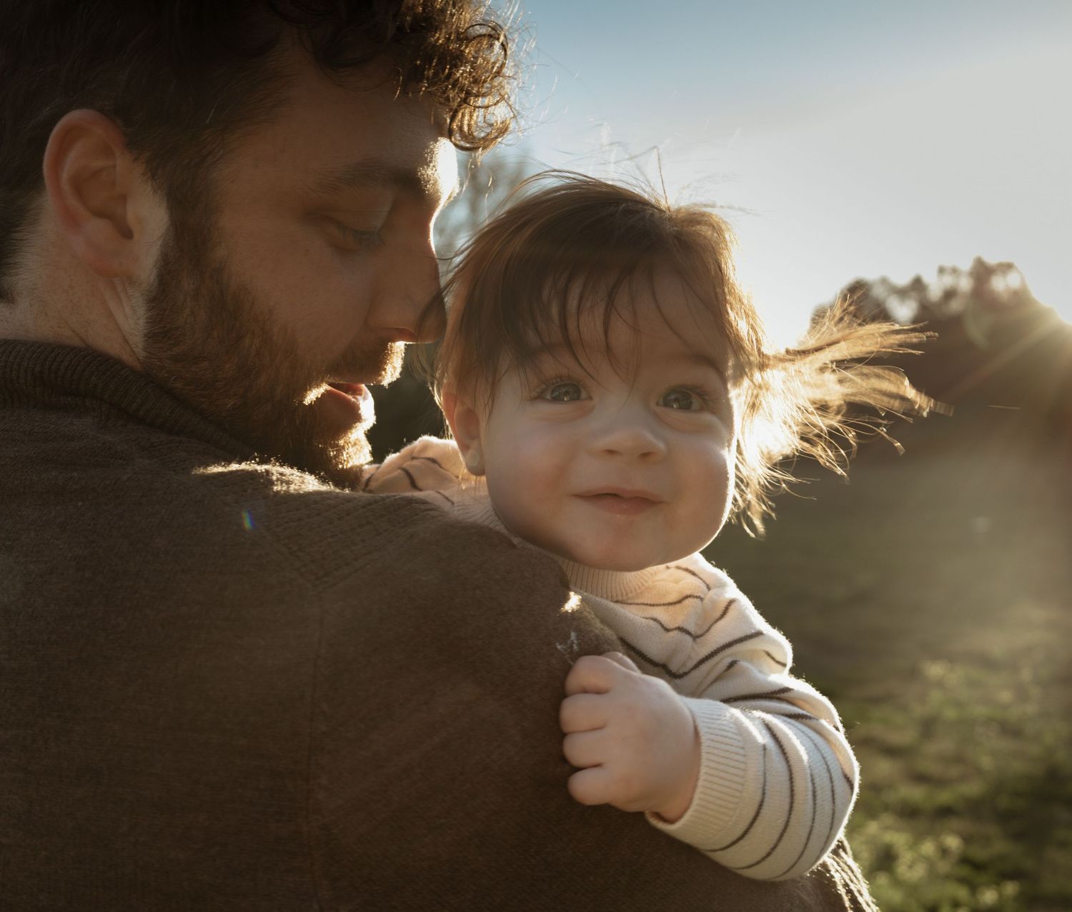 A bearded father tenderly holding his smiling baby in golden sunlight outdoors.