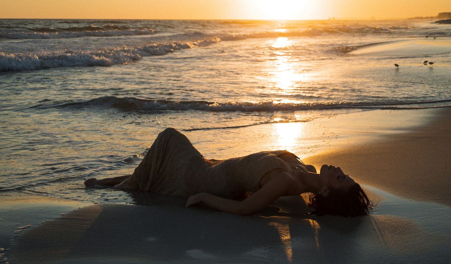 Woman in white dress lying on sandy beach at golden sunset with ocean waves.