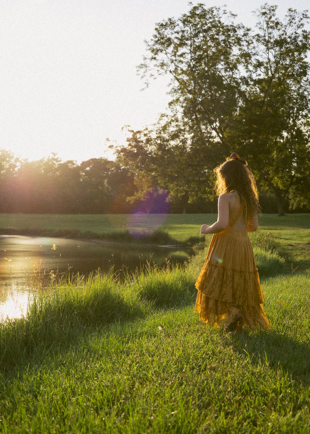 Woman in golden yellow dress walking by a sunlit pond surrounded by lush green grass and trees at sunset.