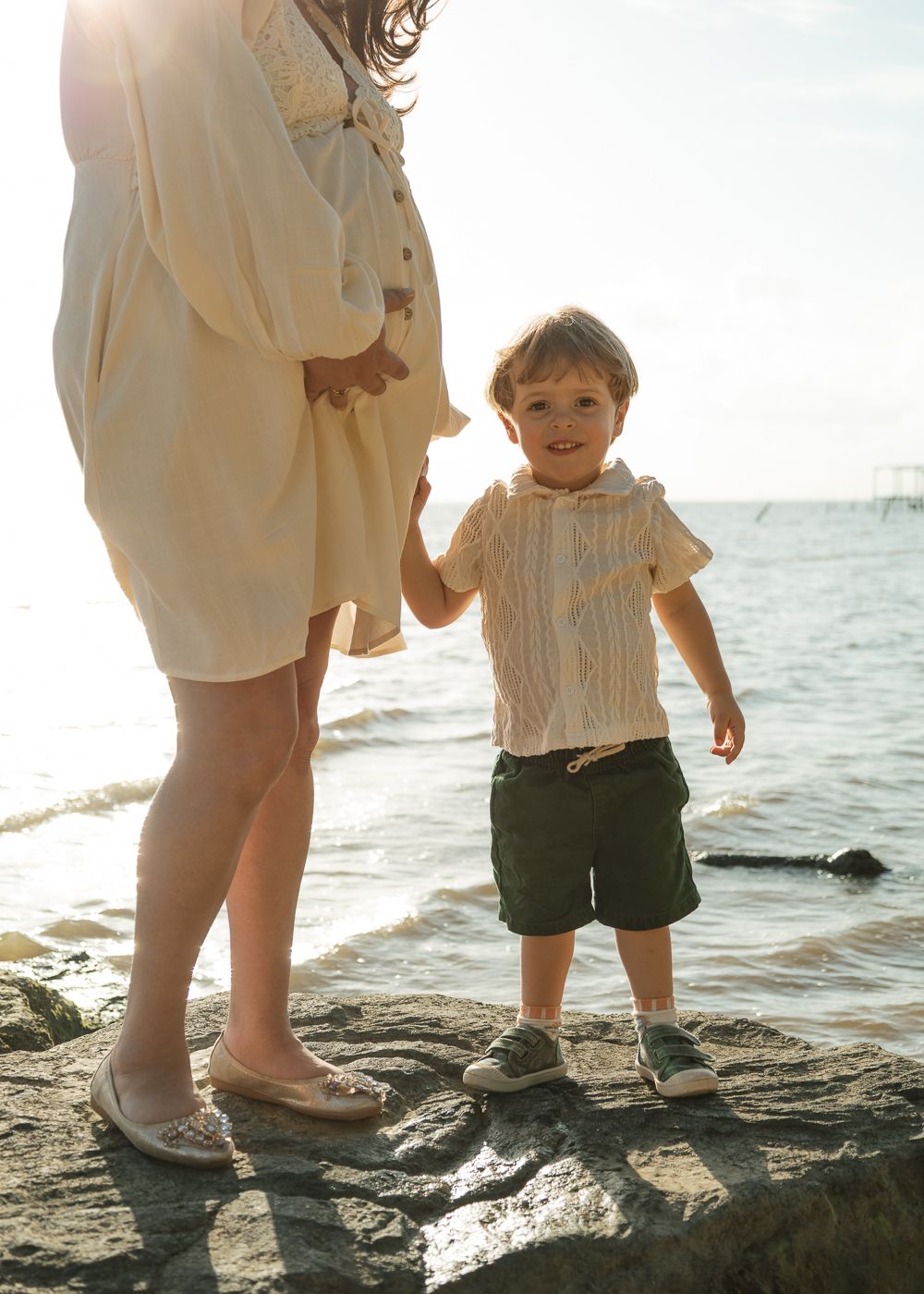 A young toddler boy in a striped shirt and green shorts holds hands with a woman in a cream dress on rocky seaside.