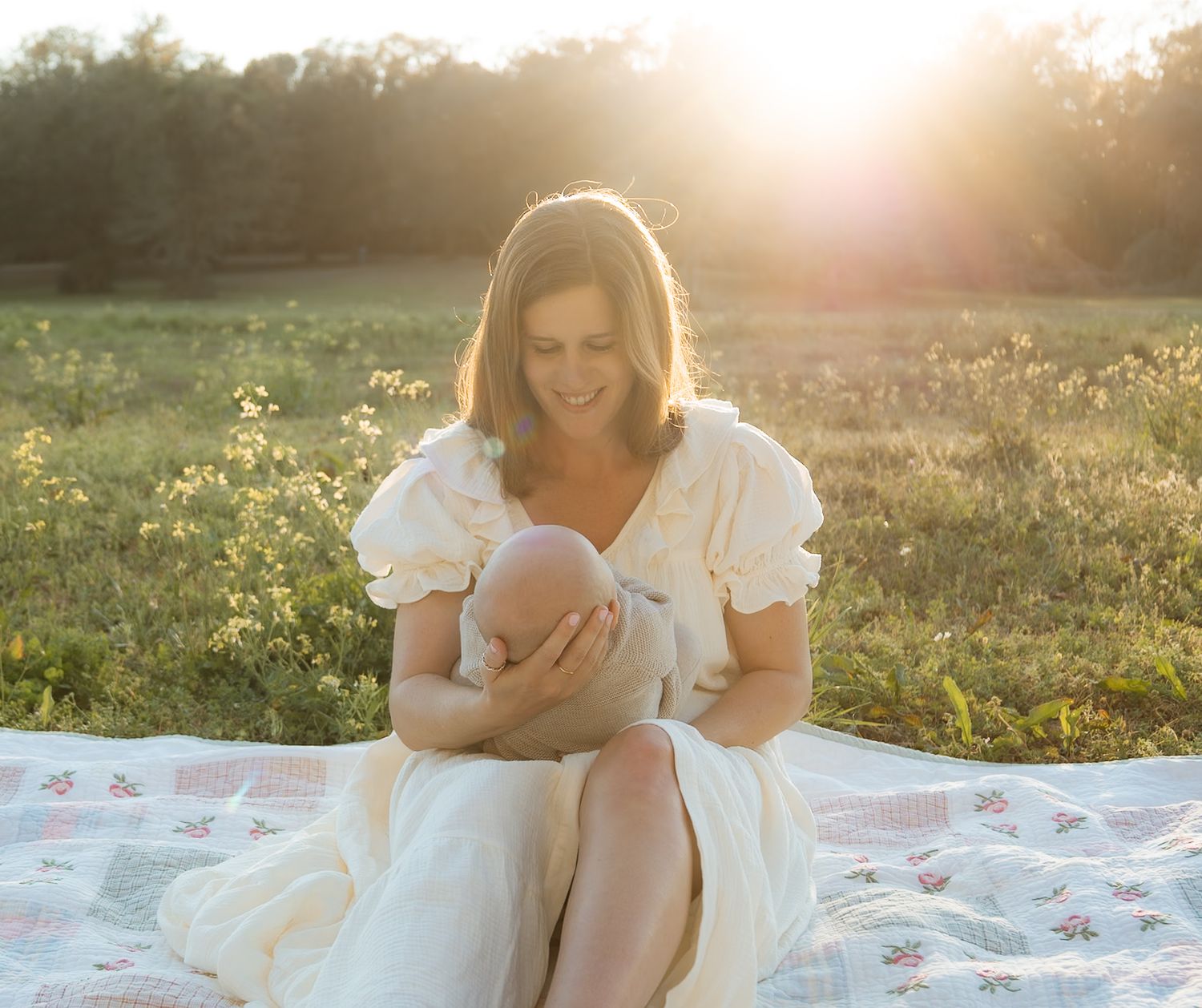 Mother in vintage cream dress sitting on quilt nursing newborn baby in golden sunset field.