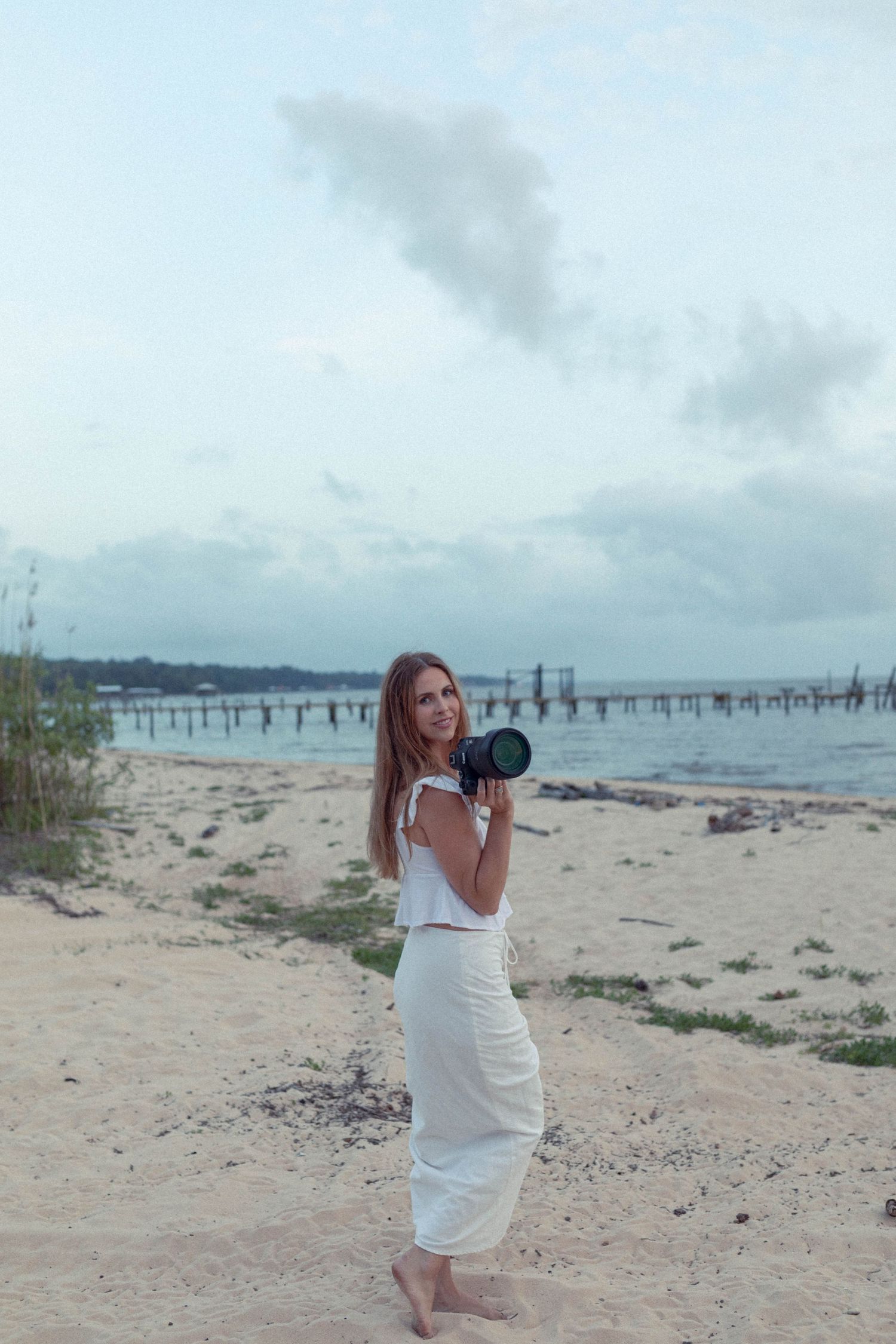 Female photographer in white dress holding camera on sandy beach at dusk.
