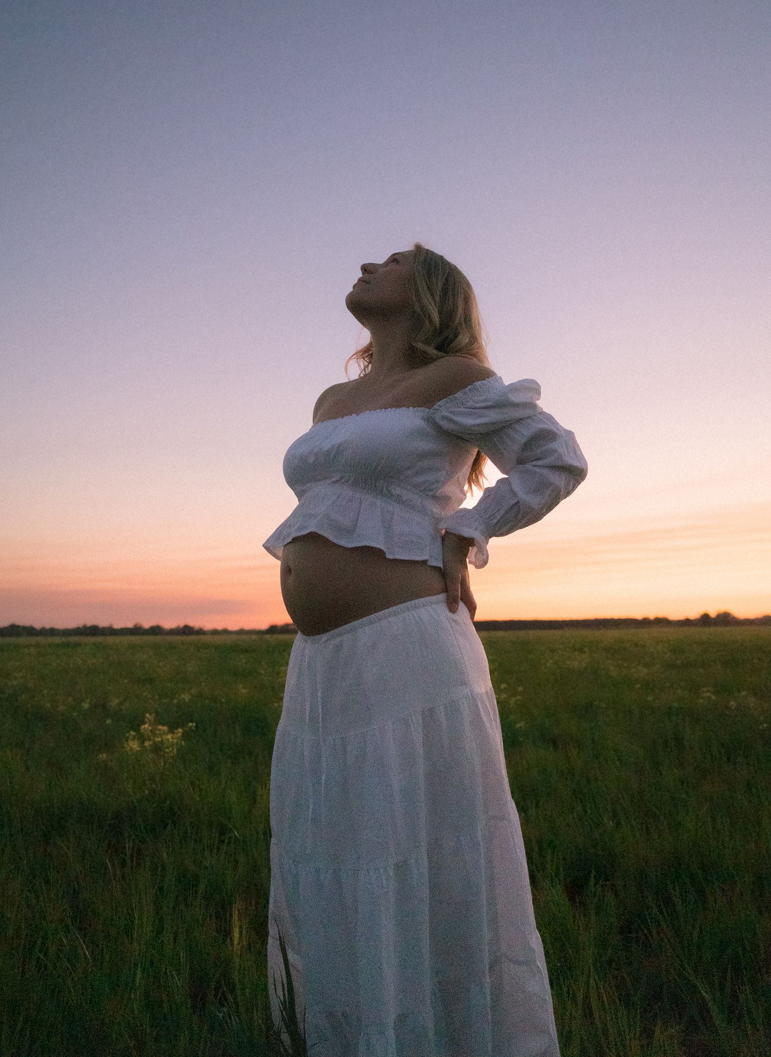 Pregnant woman in white off-shoulder crop top posing against purple twilight sky.