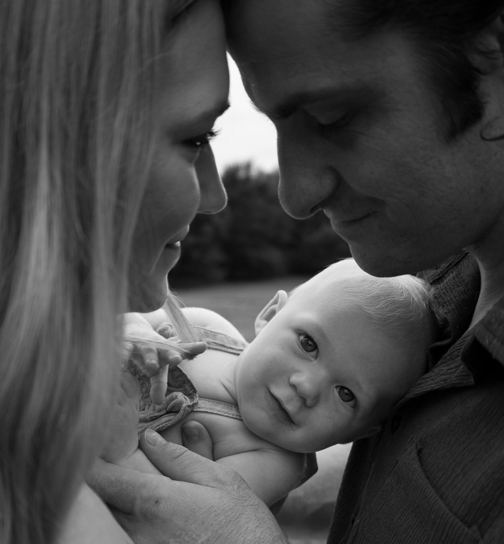Black and white photo of smiling baby held by two parents looking down lovingly in an outdoor setting.