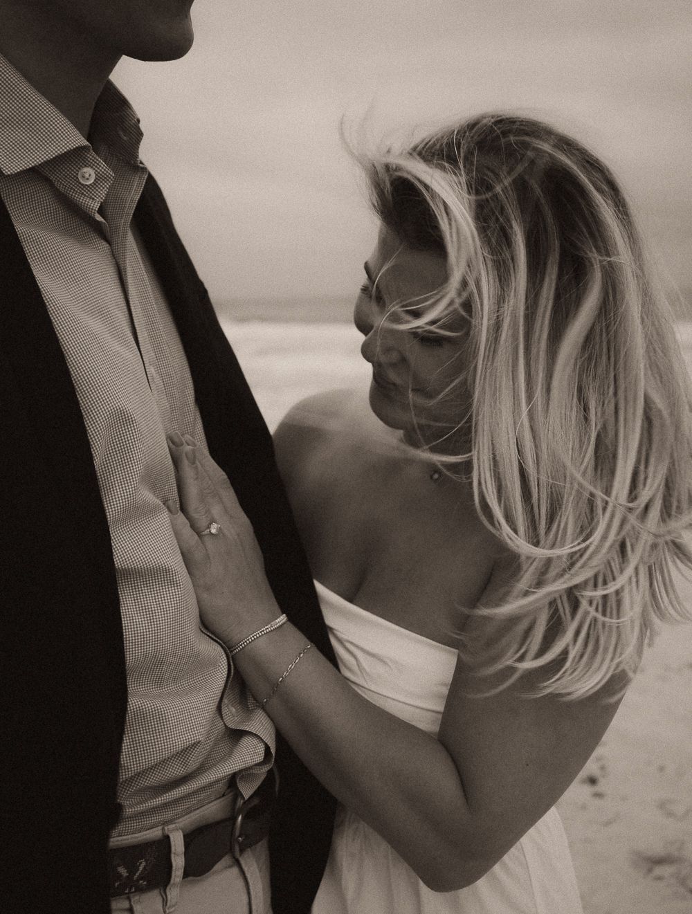 Romantic couple embracing on a beach, black and white photo, blonde woman in white dress with wind-blown hair.
