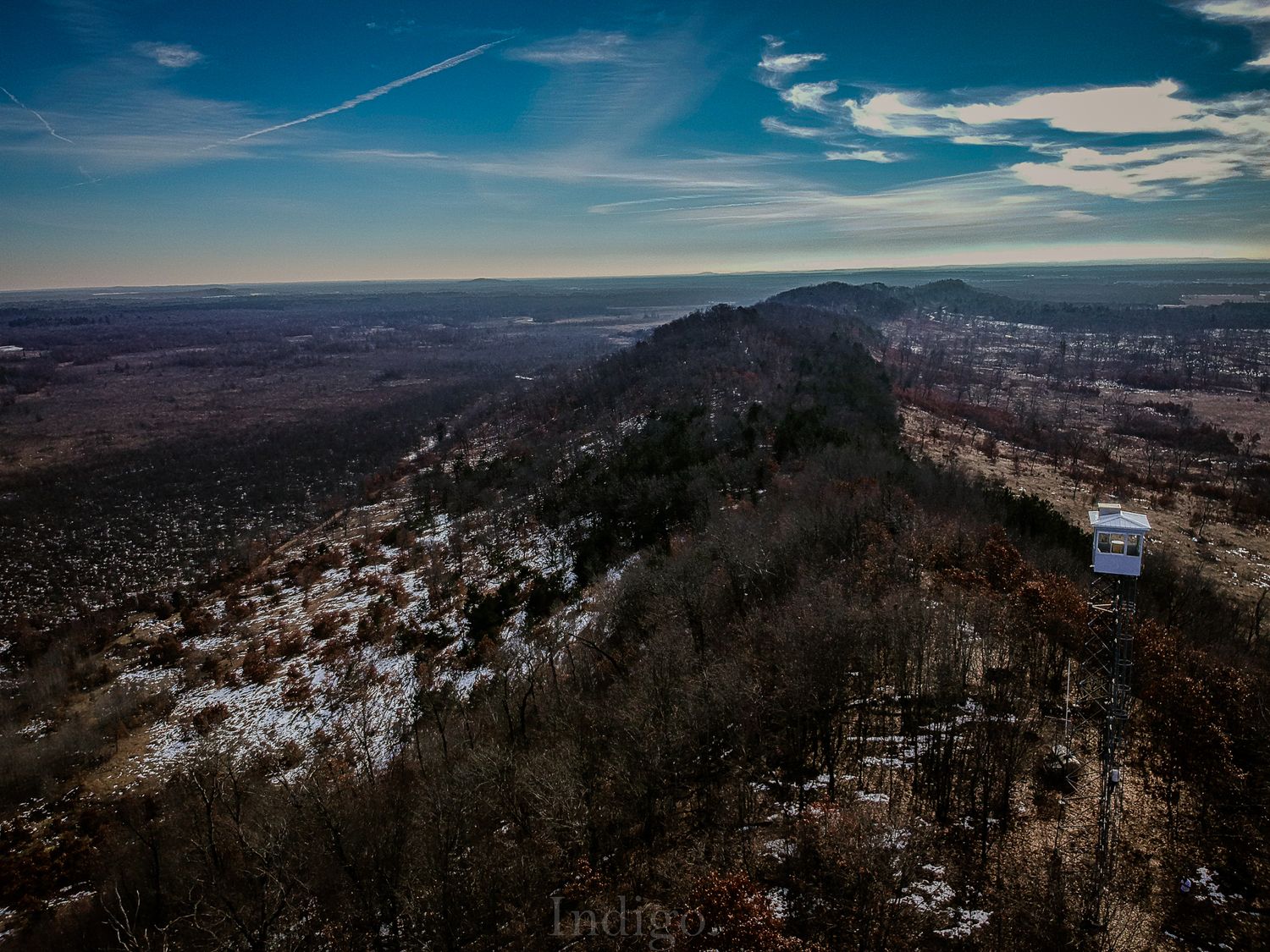 Aerial view of a snowy mountain ridge with dense forest and dramatic blue sky over a vast wilderness landscape.