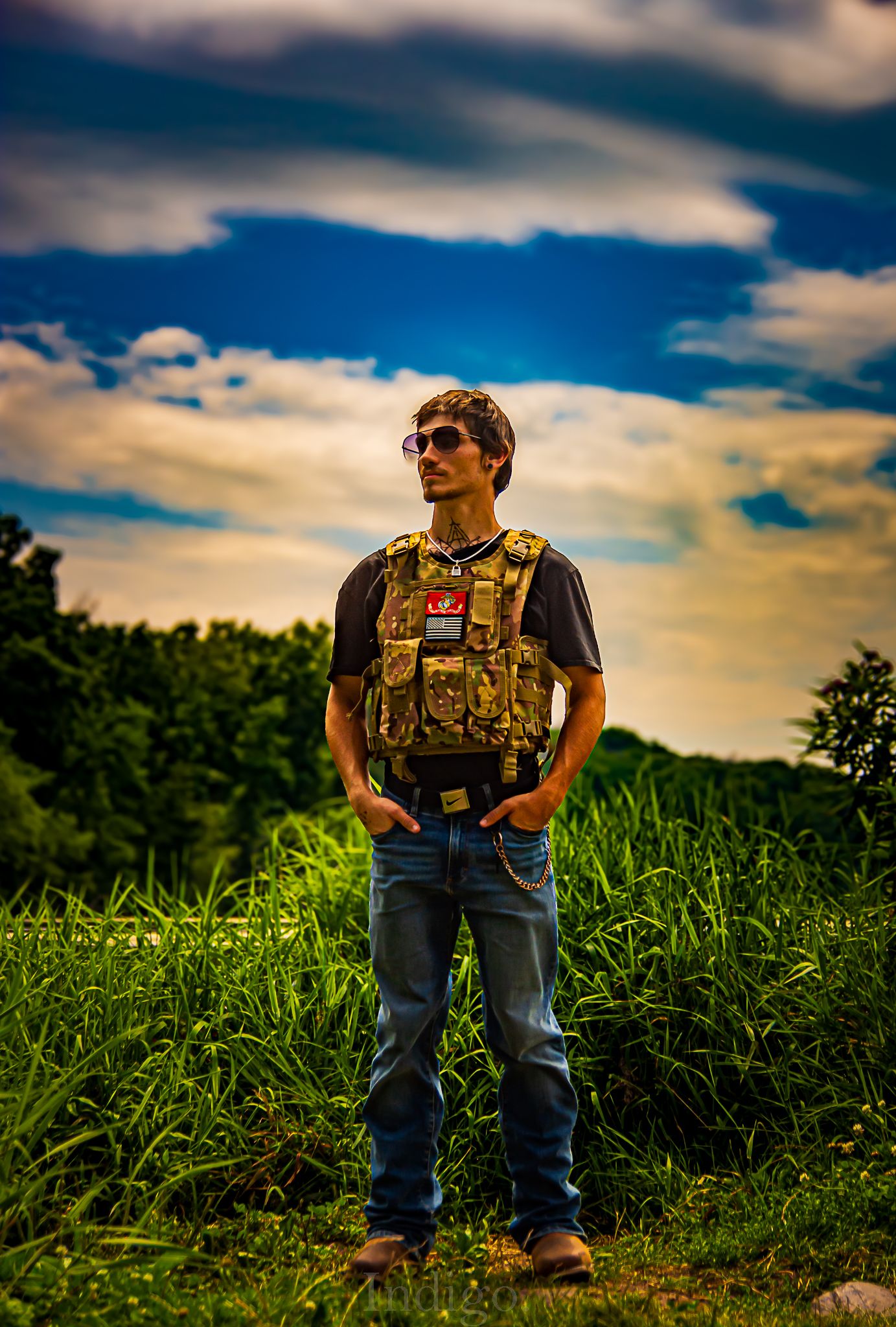 Person wearing tactical vest stands in grassy field under dramatic sunset sky.