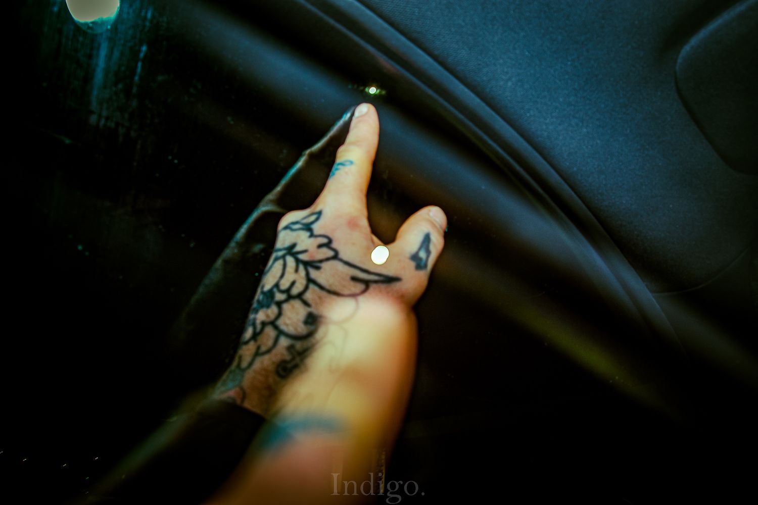 A tattooed hand with painted nails gestures against a dark background in a moody, artistic photograph.