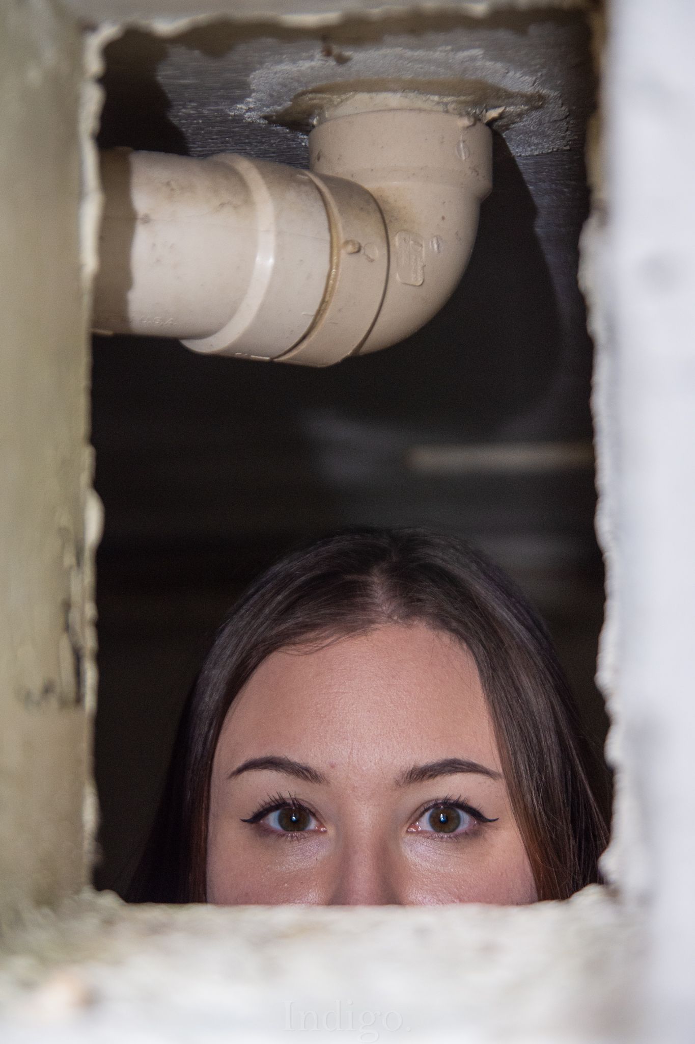 Eyes peering through a small opening in a wall with plumbing pipes visible above.