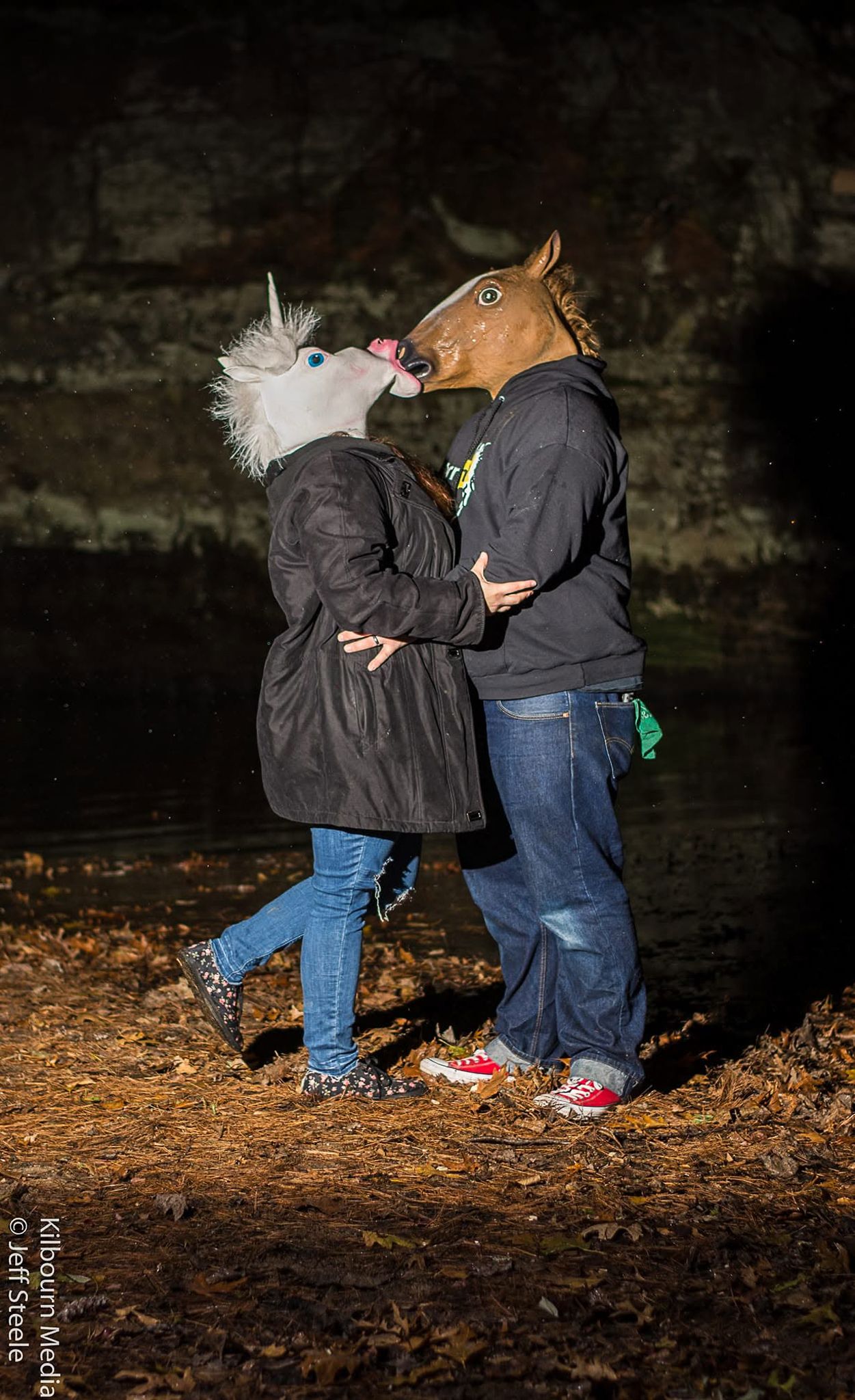 Two people in jeans and dark jackets sit together on fallen autumn leaves in a dimly lit setting.