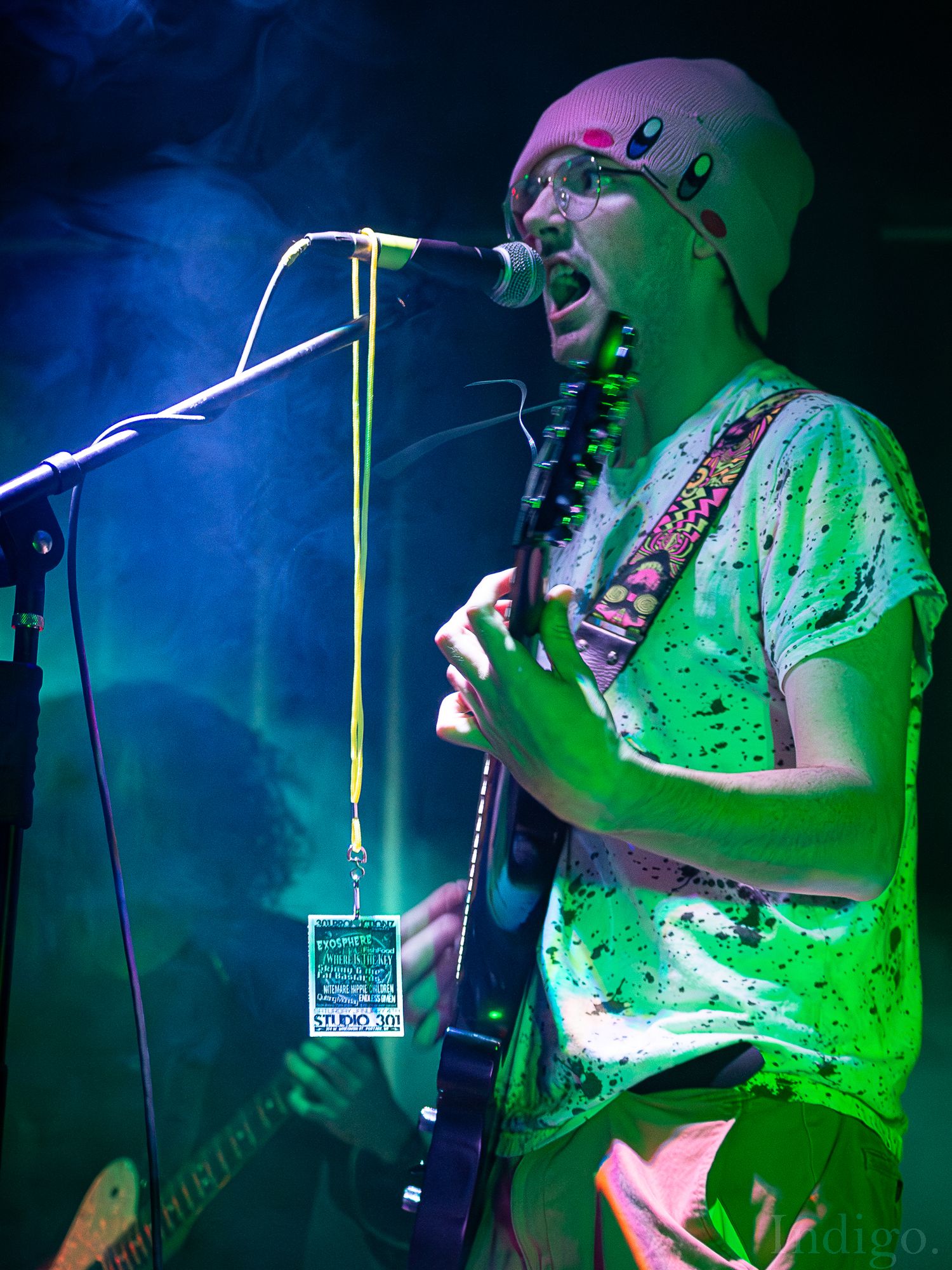 Musician bathed in green stage lights performing with instruments and equipment visible on stage.