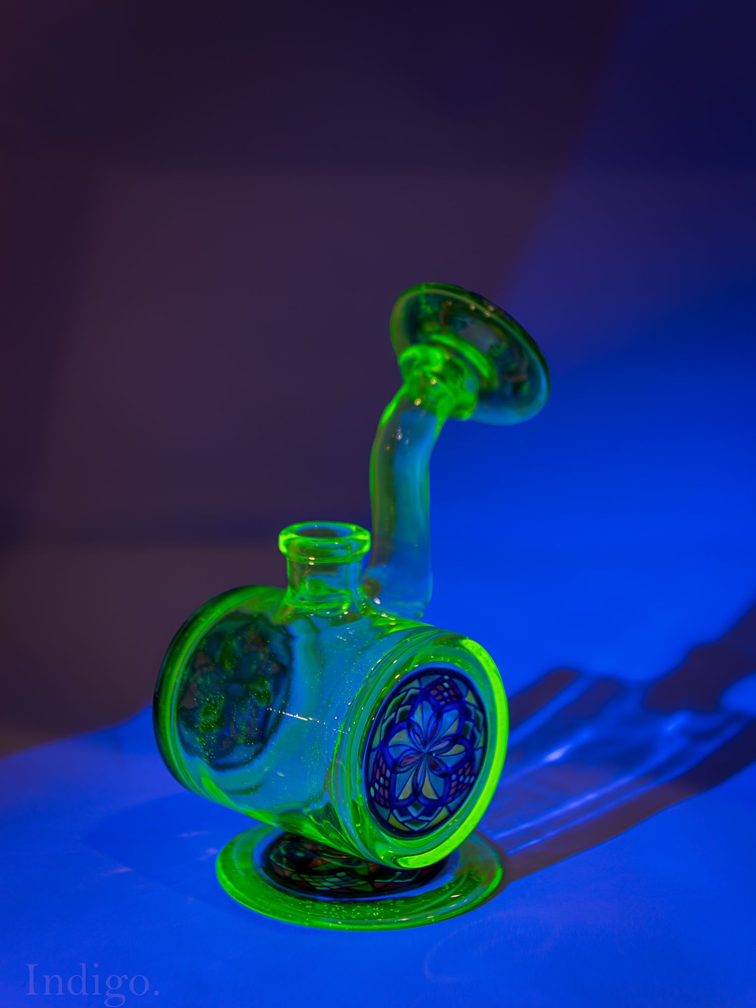 Glass smoking device illuminated by blue and green lighting in a dark setting.