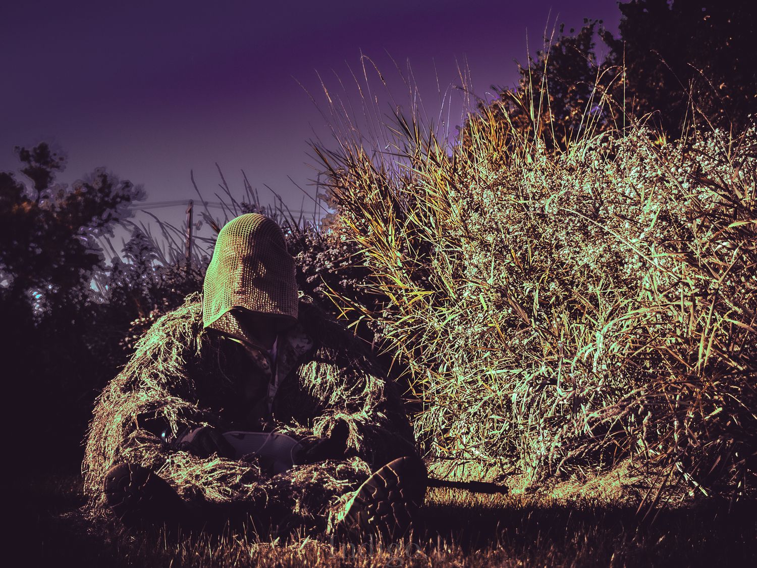 A camouflaged figure crouches in tall grass at night with an eerie green glow illuminating the scene.
