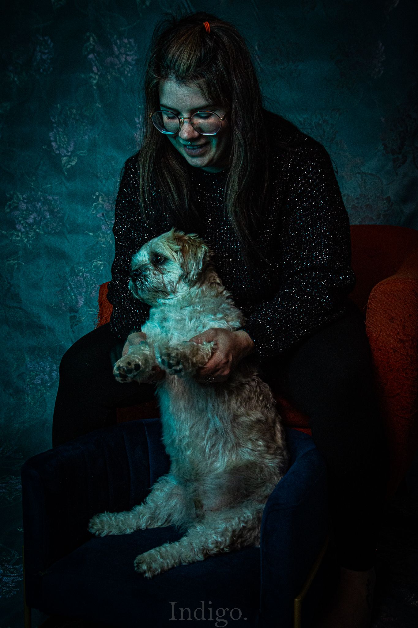 A person in dark clothing sits with a small white fluffy dog against a moody teal backdrop in dramatic lighting.