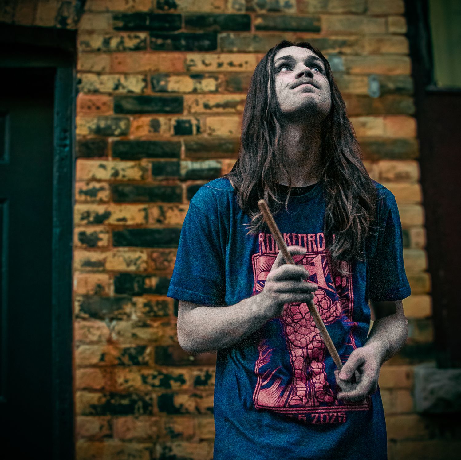 Long-haired musician in blue shirt looking upward against brick wall backdrop.