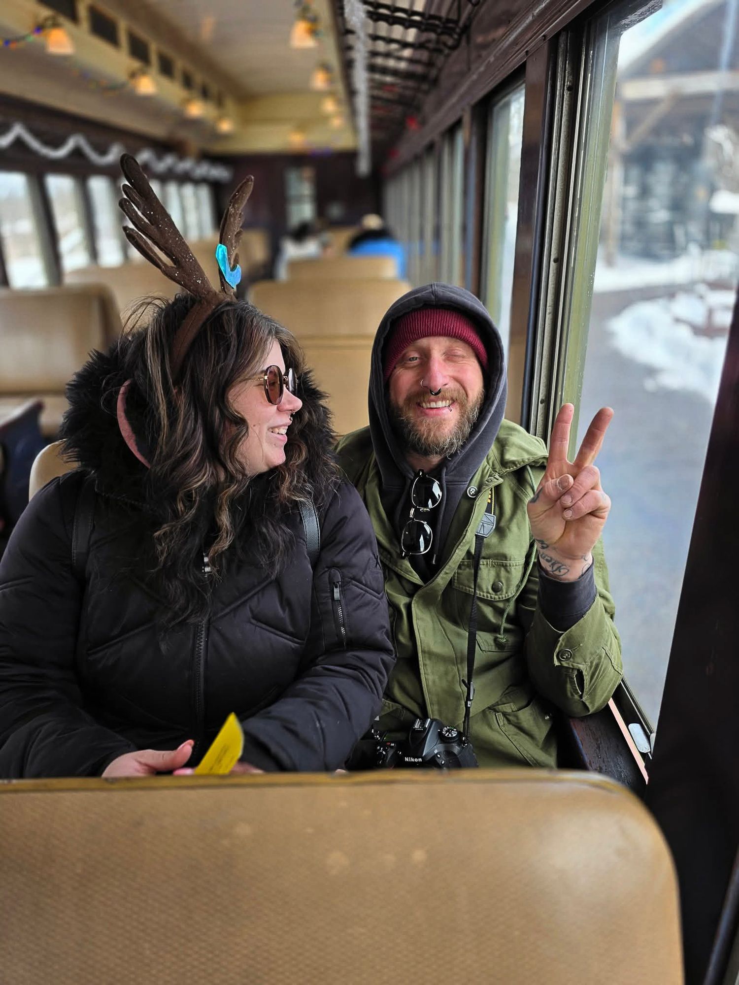 Two passengers share a laugh while riding on a train, sitting on tan seats next to a window on a winter day.