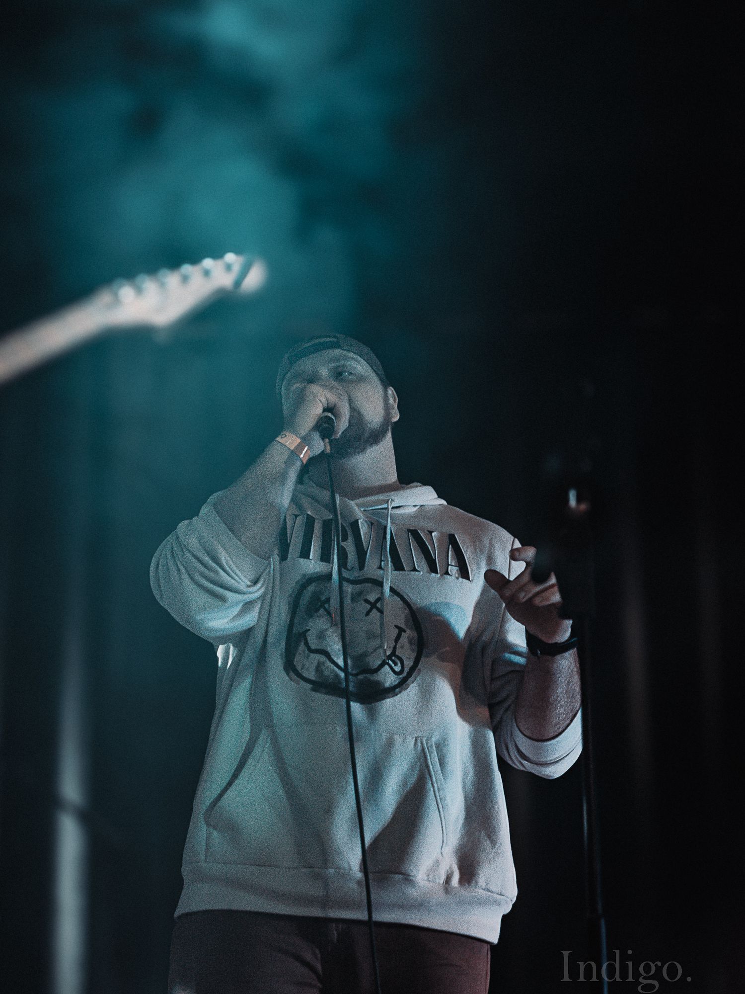 Performer in white hoodie singing into microphone with turquoise stage lighting.