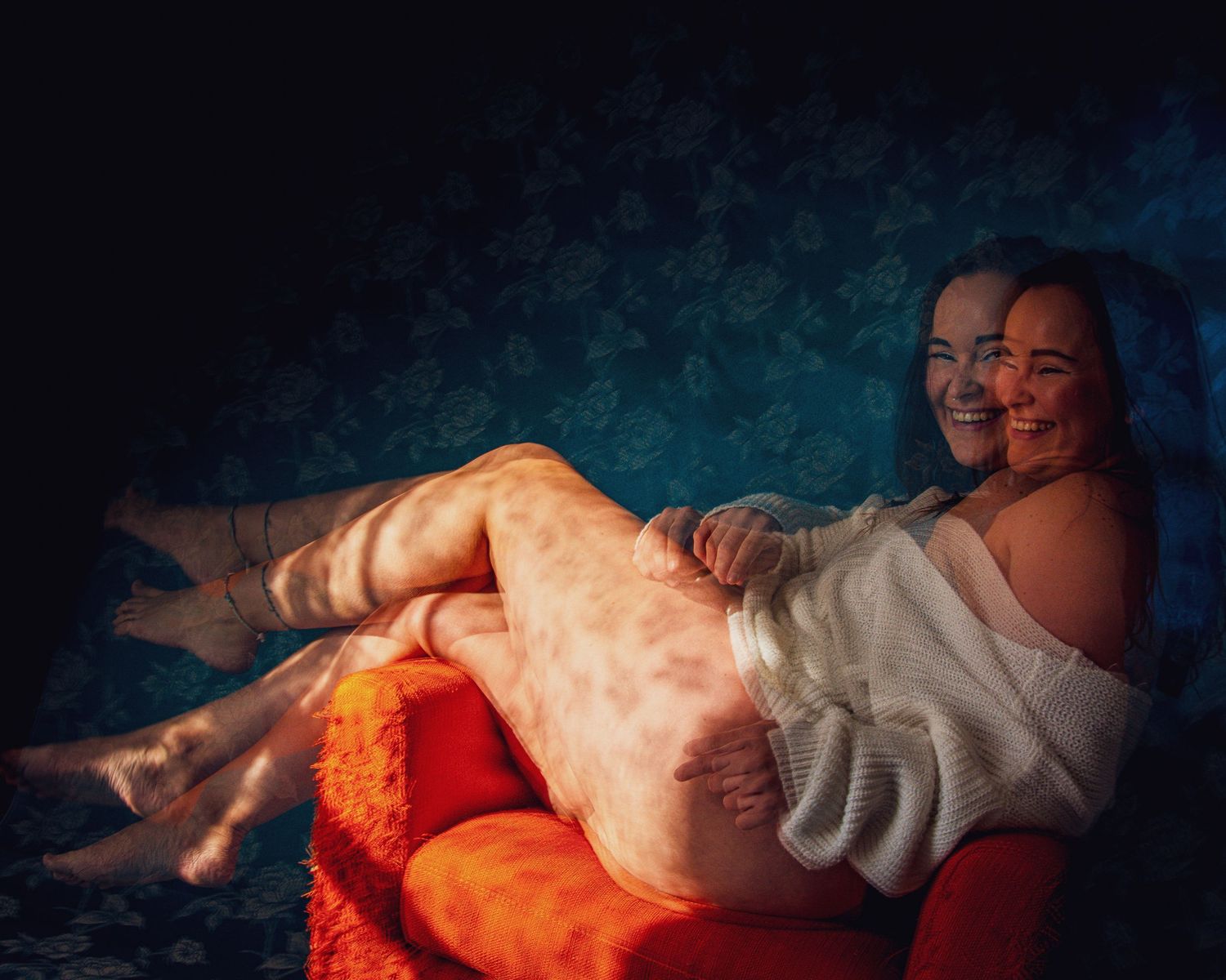Two smiling women relaxing together on a vibrant red-orange chair against a blue floral wallpaper background.