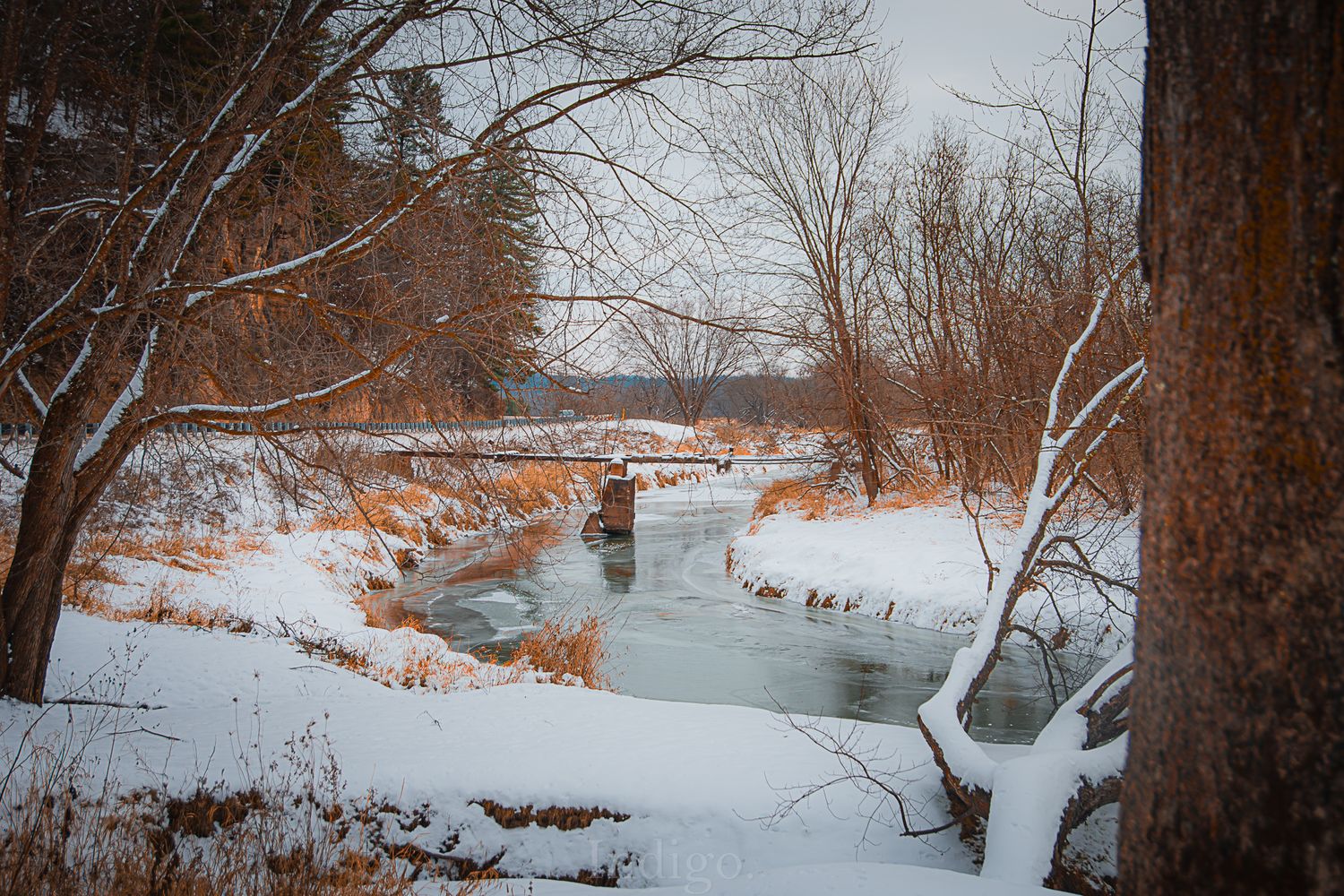 Snow-covered riverbank in winter with bare trees and a peaceful stream winding through the landscape.