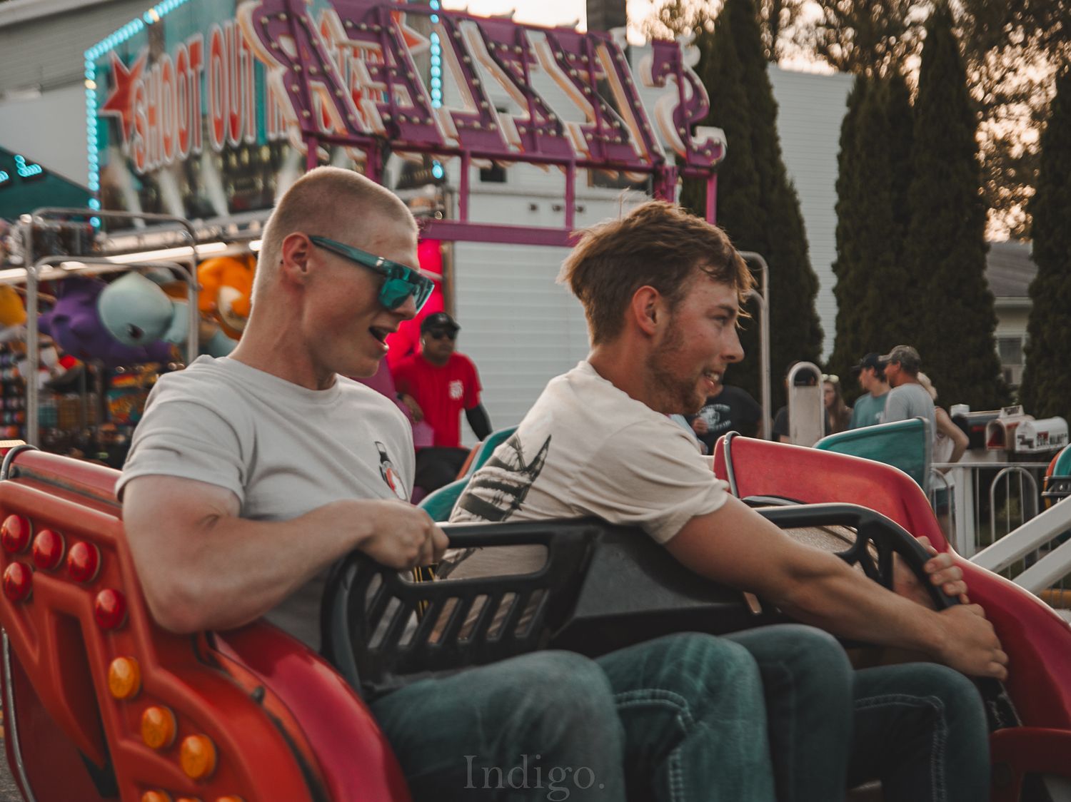 People sit in a row of red carnival ride seats at dusk during an outdoor fair or amusement park.
