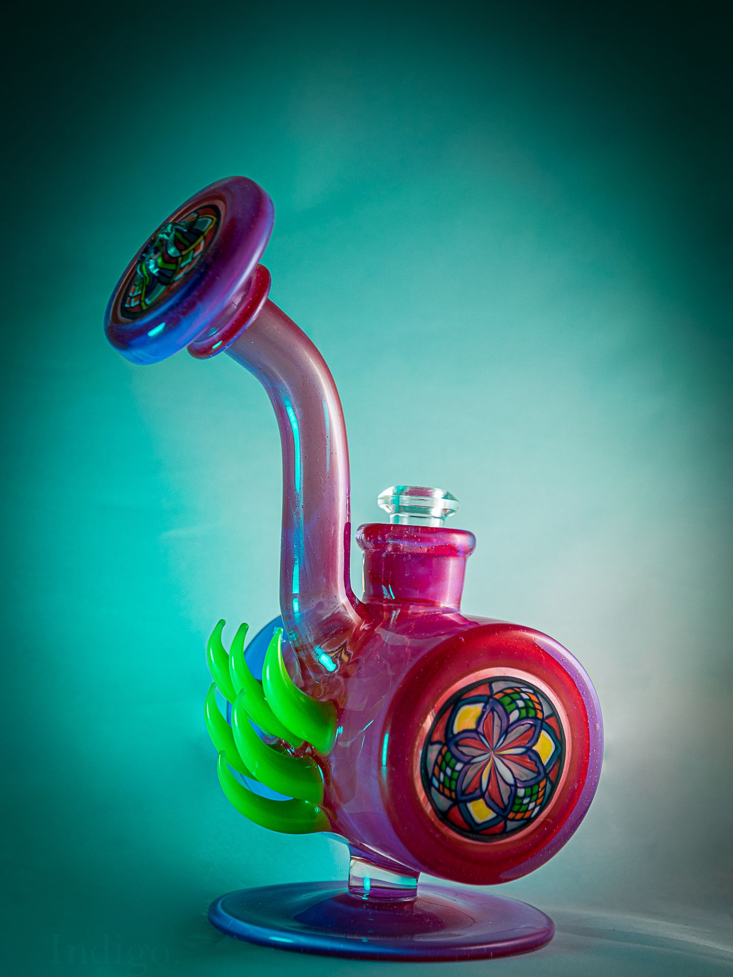 Artistic glass smoking device with pink and green accents on teal backdrop.