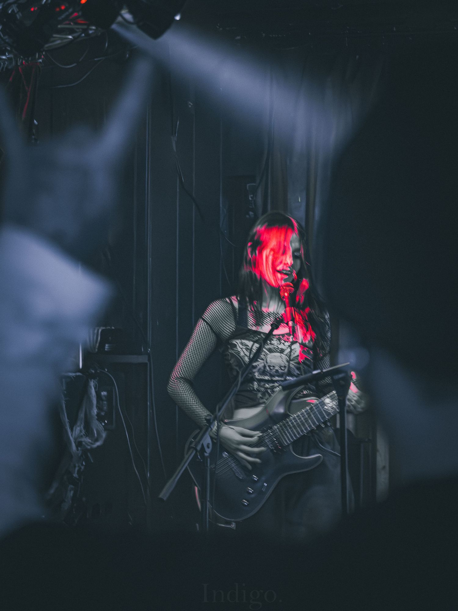 Concert performer playing guitar on dark stage with dramatic red lighting and atmospheric smoke effects.