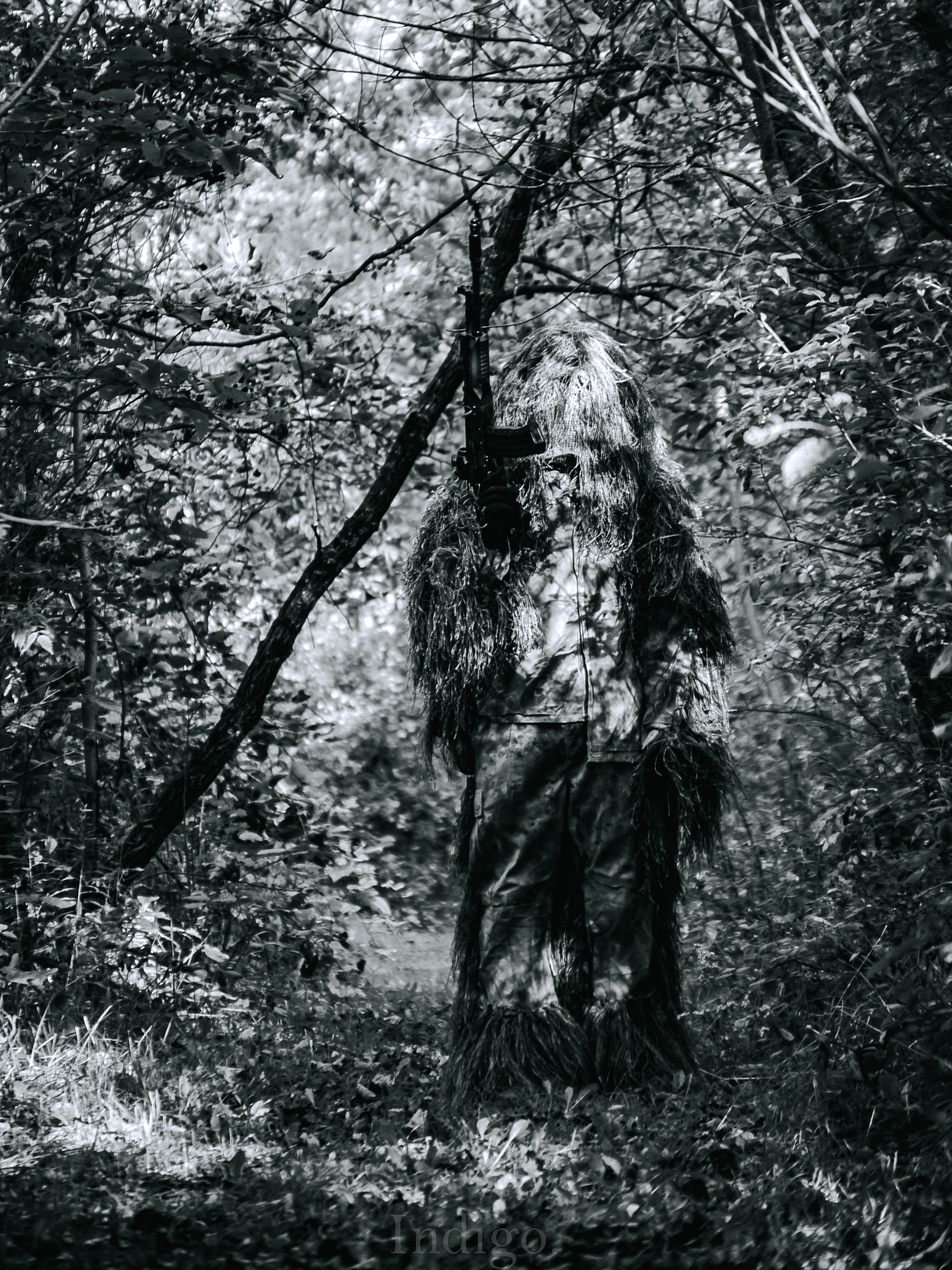 A spooky black and white photo of a haunting forest creature with long hair and tattered clothes standing among trees.