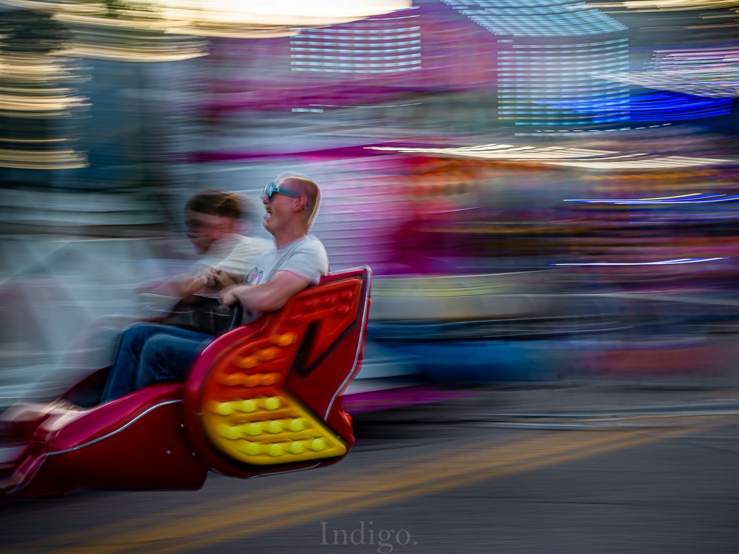 Motion-blurred carnival rides at dusk create colorful light trails against a darkening sky.