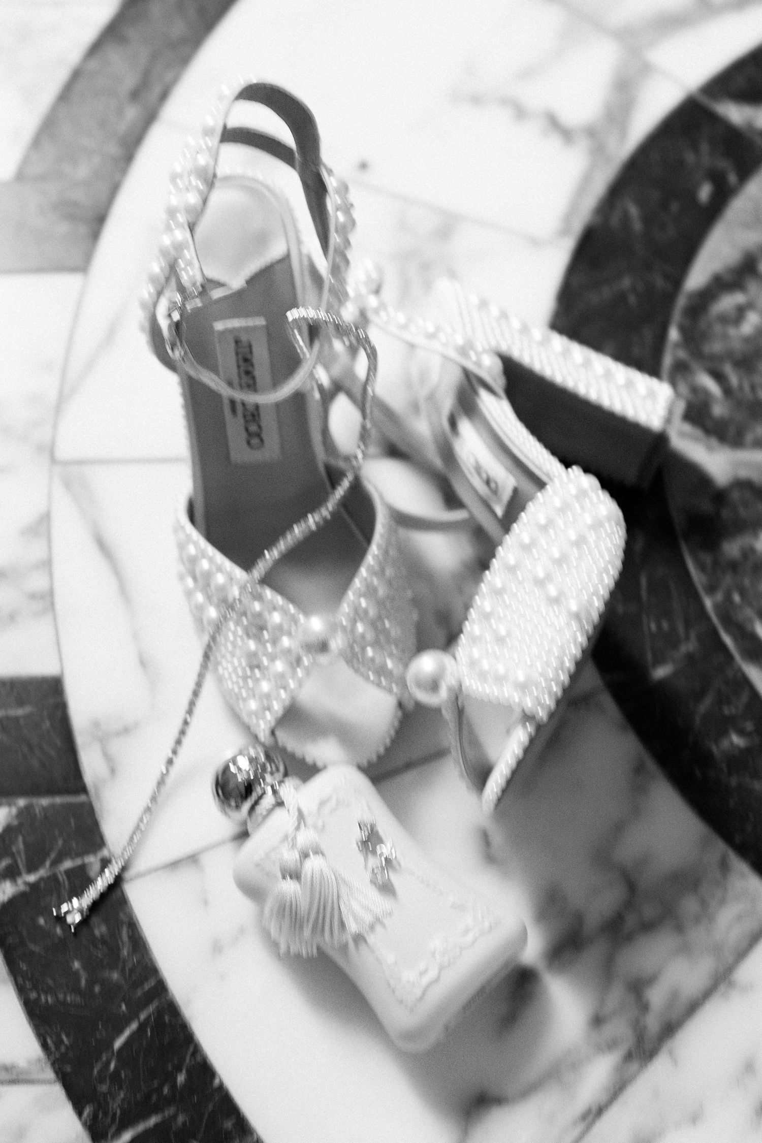 Black and white photo of pearl-embellished heeled sandals with a mini tassel bag on marble surface.