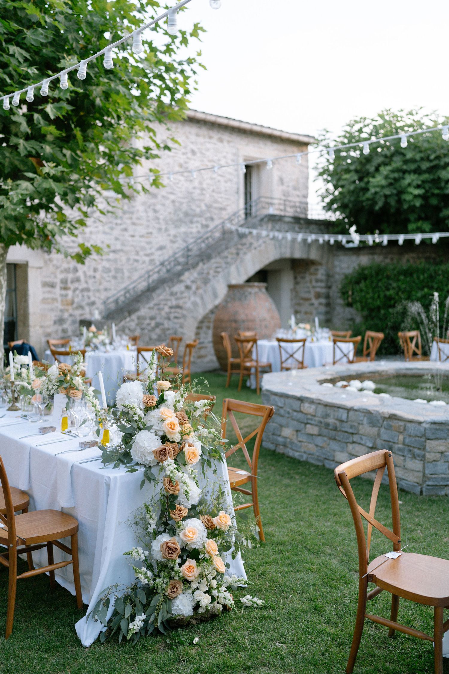 Outdoor wedding reception setup with white tablecloths and wooden chairs against a stone wall backdrop.