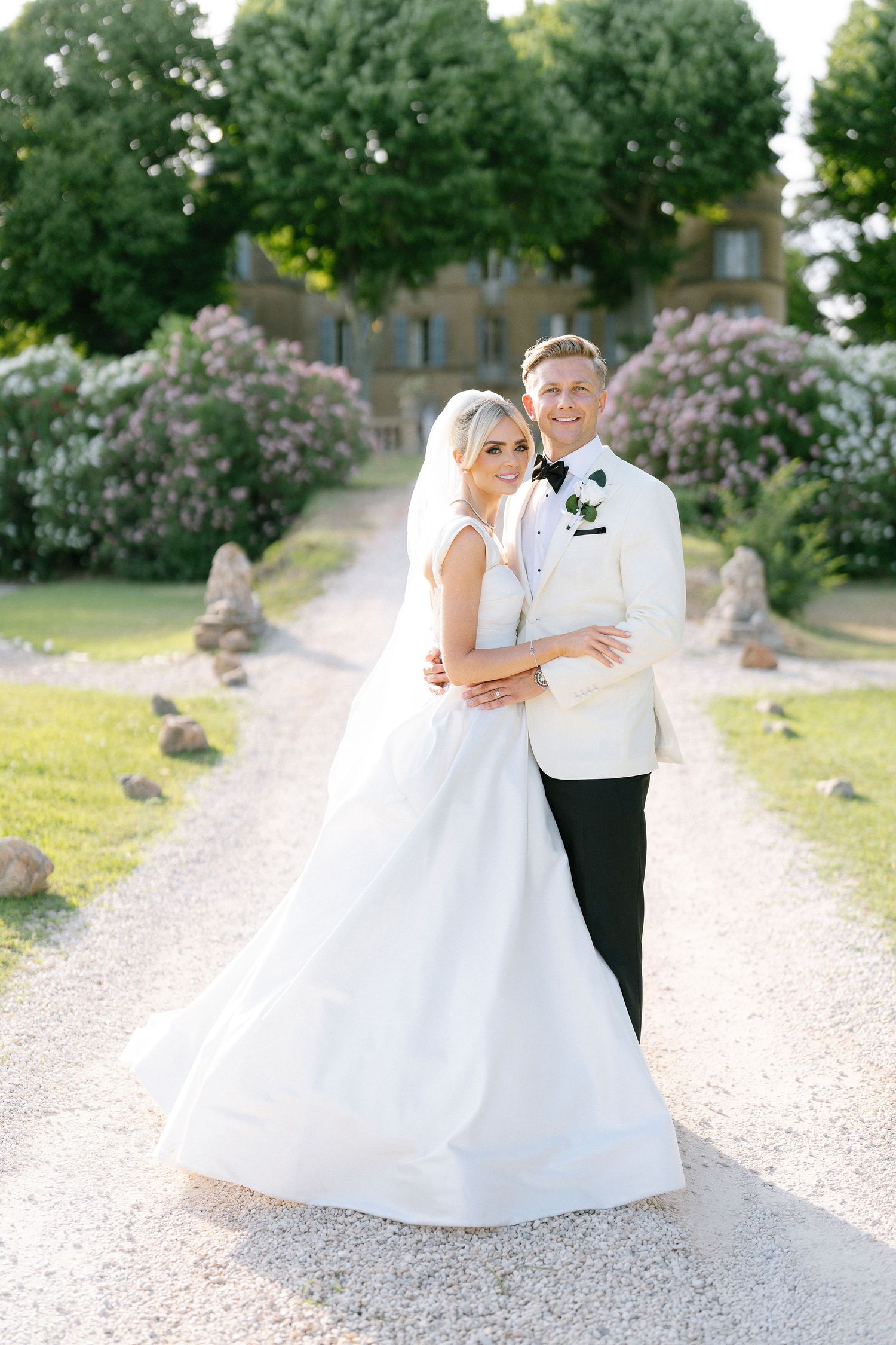 Bride and groom embrace on gravel path before elegant chateau surrounded by blooming gardens on their wedding day.