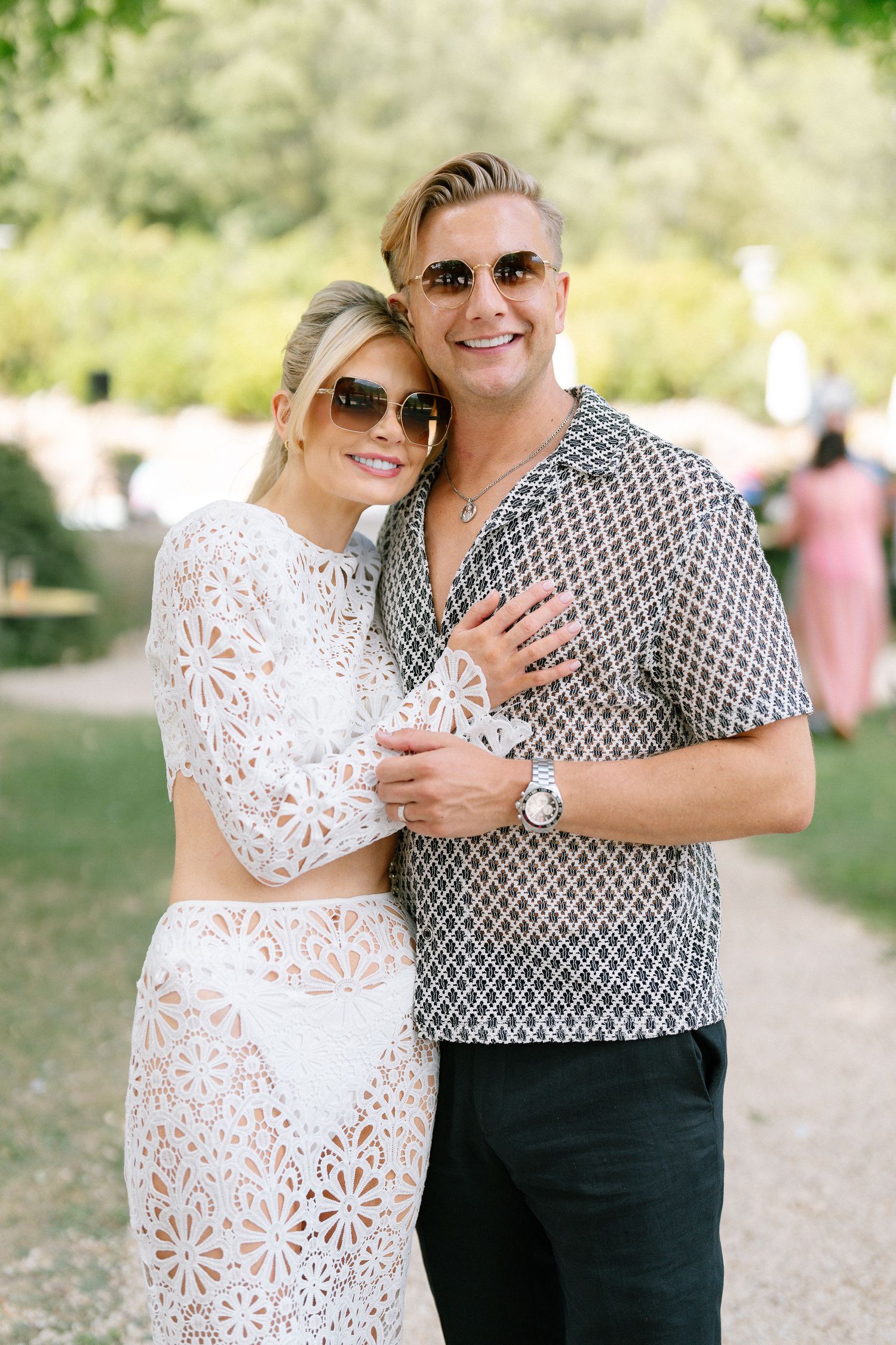Smiling couple in sunglasses pose outdoors at a wedding, woman in white lace co-ord and man in patterned shirt.