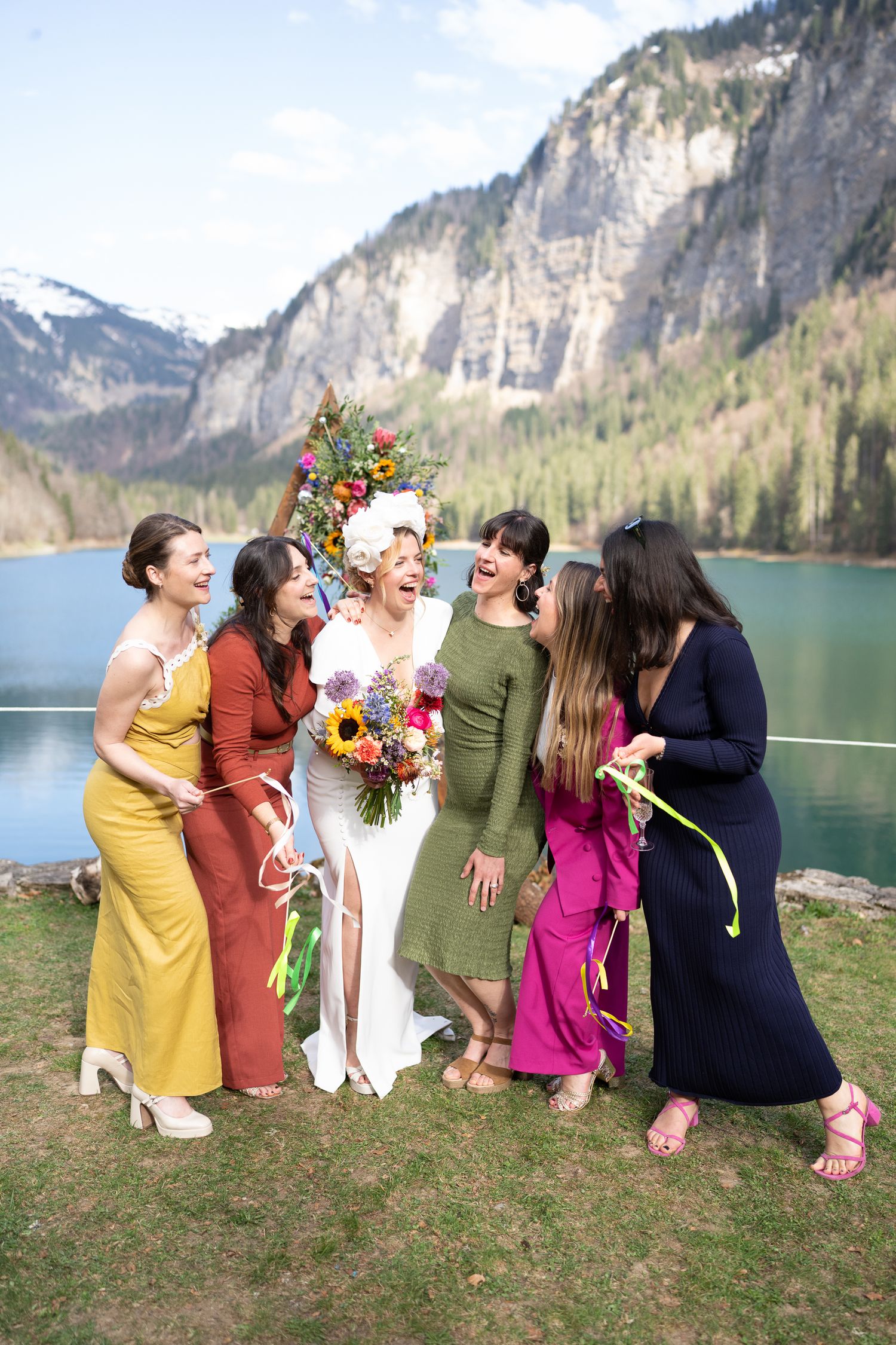Group of friends in colorful dresses laughing together by a scenic mountain lake with spring flowers.