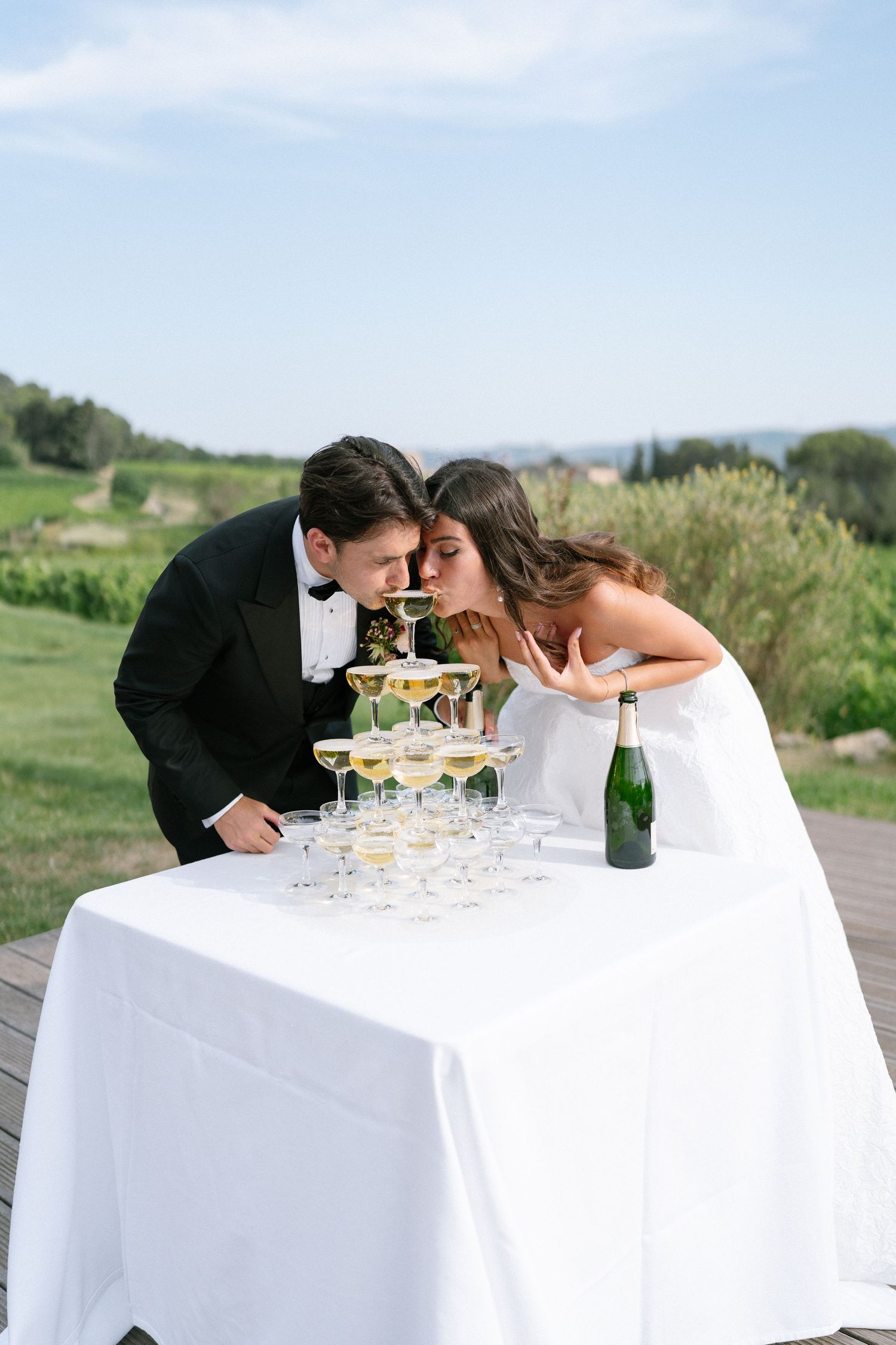 Couple playfully drinking from champagne tower setup on outdoor table.