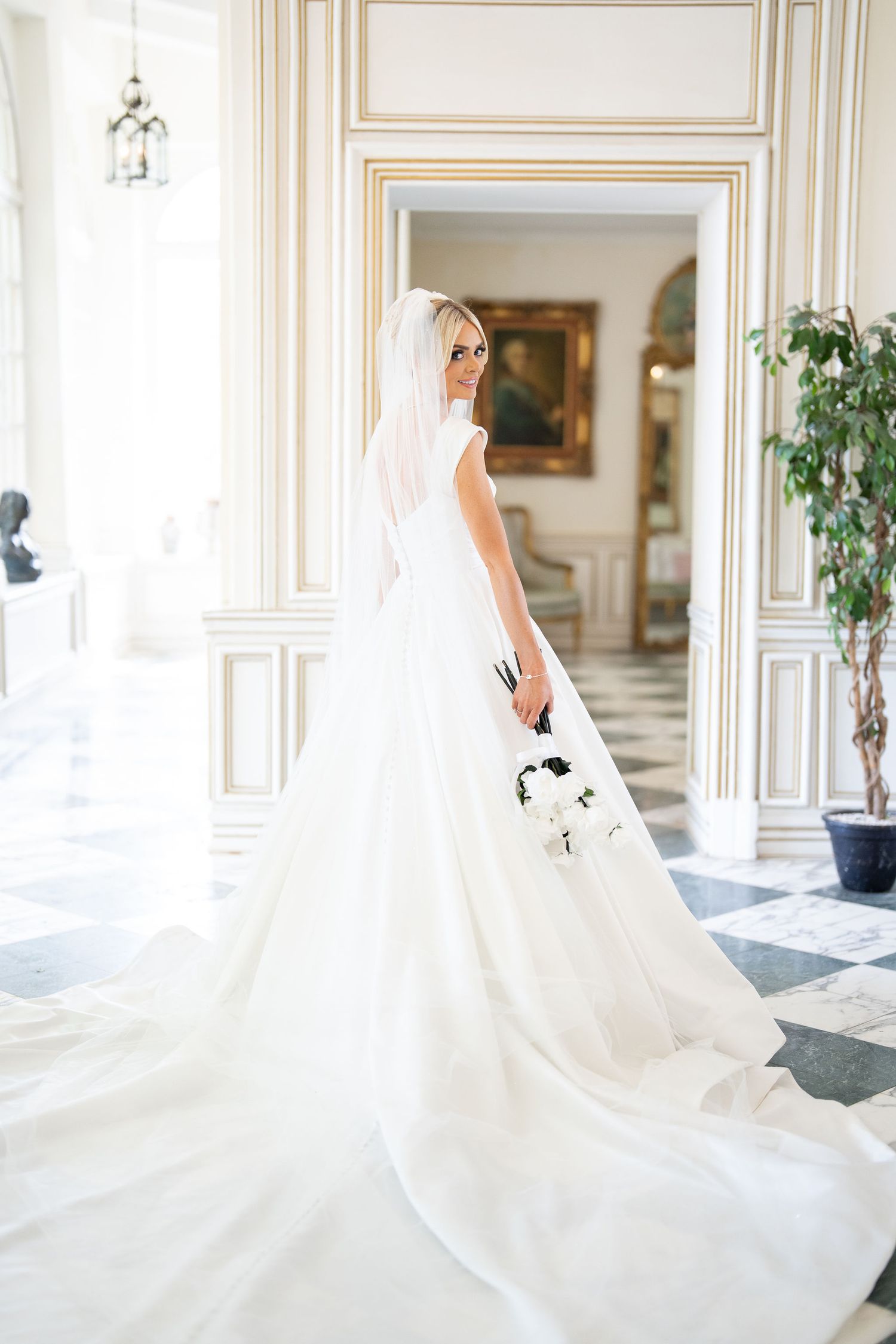 Elegant bride in a white gown with long train and veil holds bouquet in a grand, ornate hall.