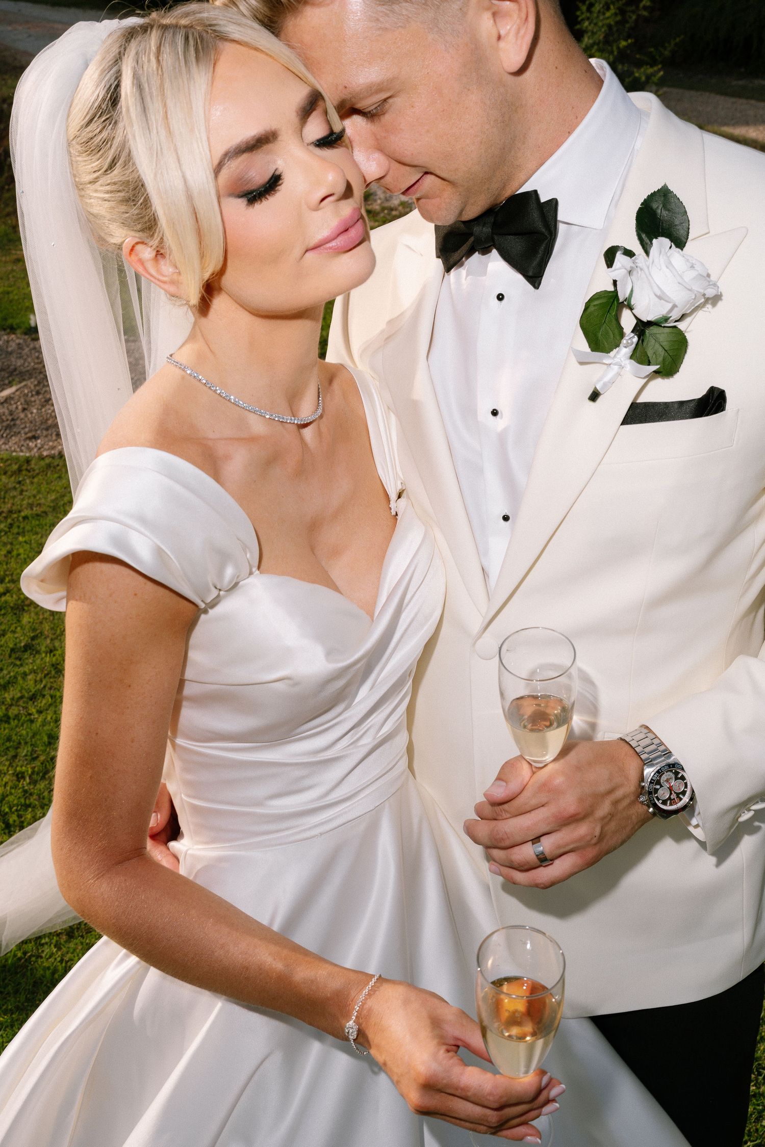 Bride and groom in white wedding attire share an intimate moment outdoors, holding champagne glasses.