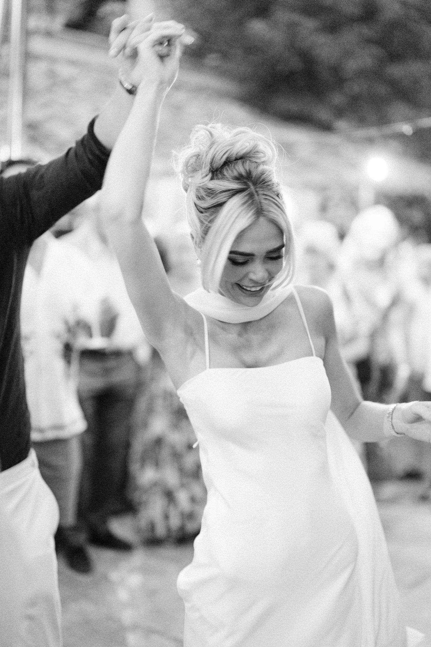 Black and white photo of a joyful bride in a white slip dress dancing with raised arms at her wedding reception.