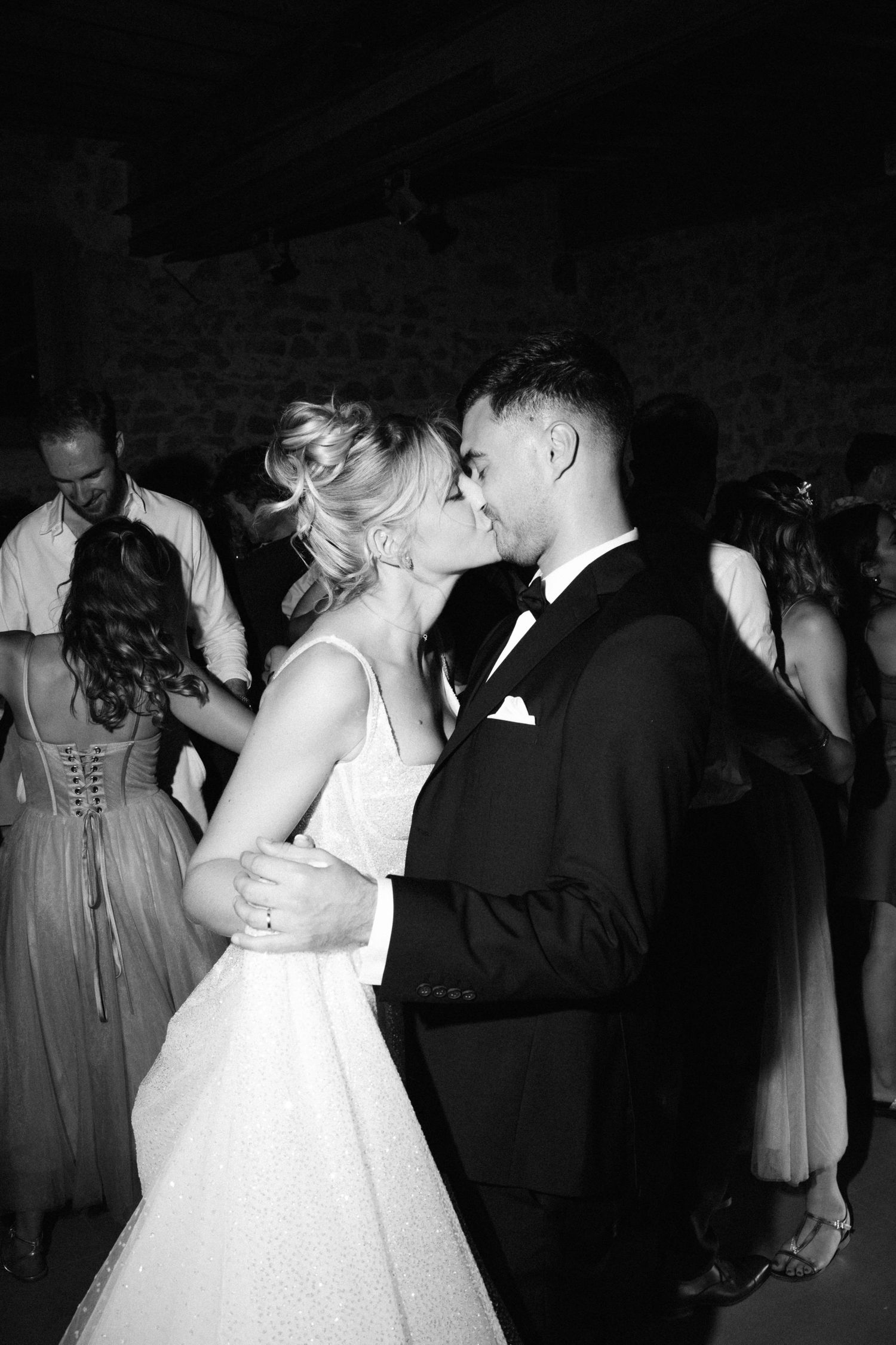 Wedding couples dance together in formal attire during a dimly lit reception.