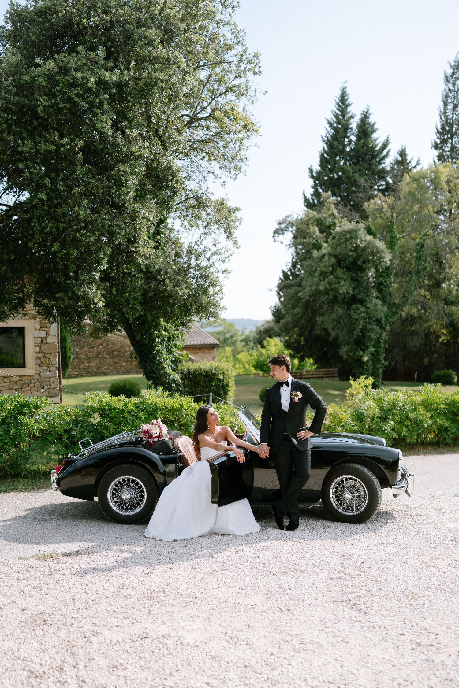 Sequence of wedding poses with vintage black sports car on gravel driveway.