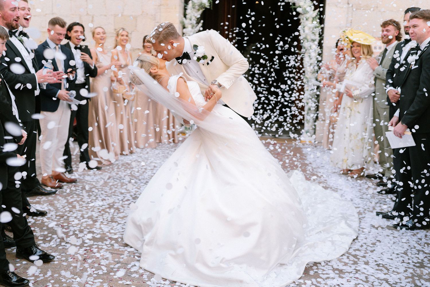 Bride and groom share romantic dip kiss surrounded by cheering guests throwing white confetti at wedding.